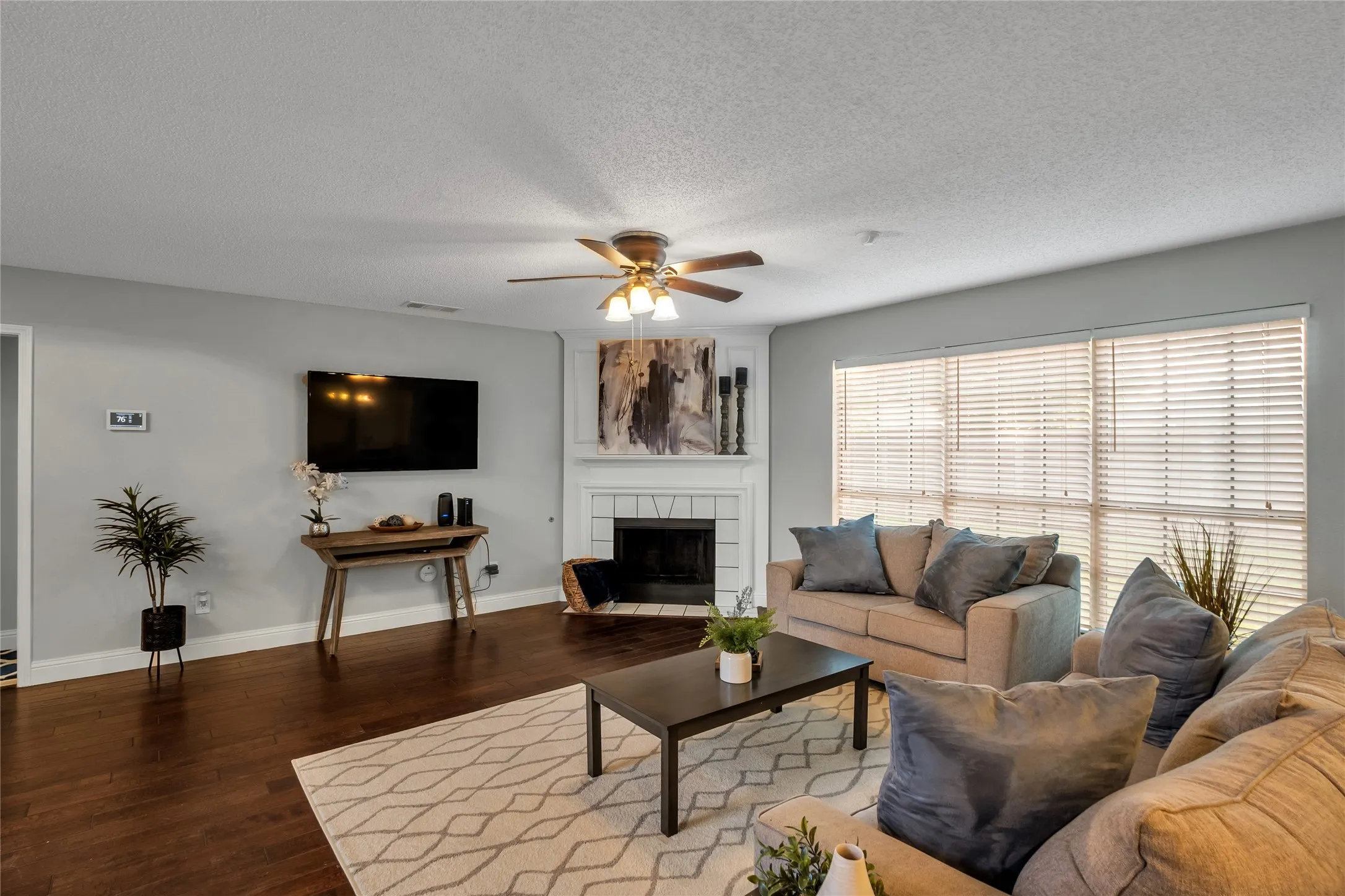 Hardwood flooring, fireplace with white mantel, ceiling fan with integrated lighting, multiple windows with blinds, and light-toned walls