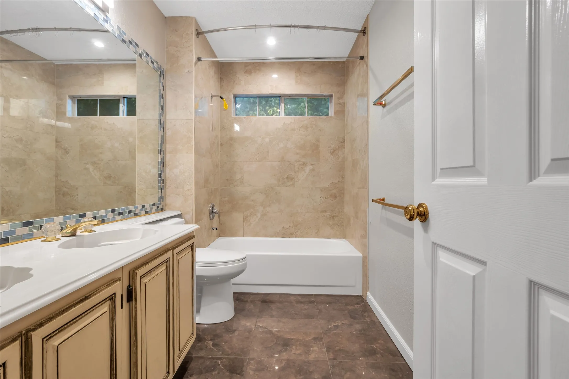 Bathroom featuring a single vanity with an integrated sink, a rectangular mirror with mosaic tile trim, a tub/shower combination with tile surround, and brown-toned tile flooring