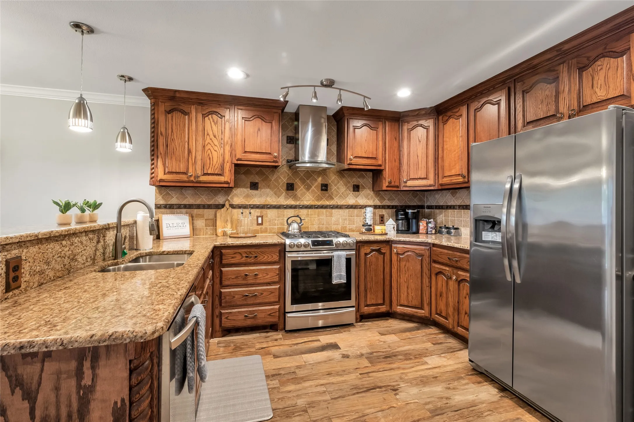 Gourmet kitchen featuring rich wood cabinetry, granite countertops, stainless steel appliances including a side-by-side refrigerator and gas range, pendant lighting, and warm wood-finish flooring throughout the open layout