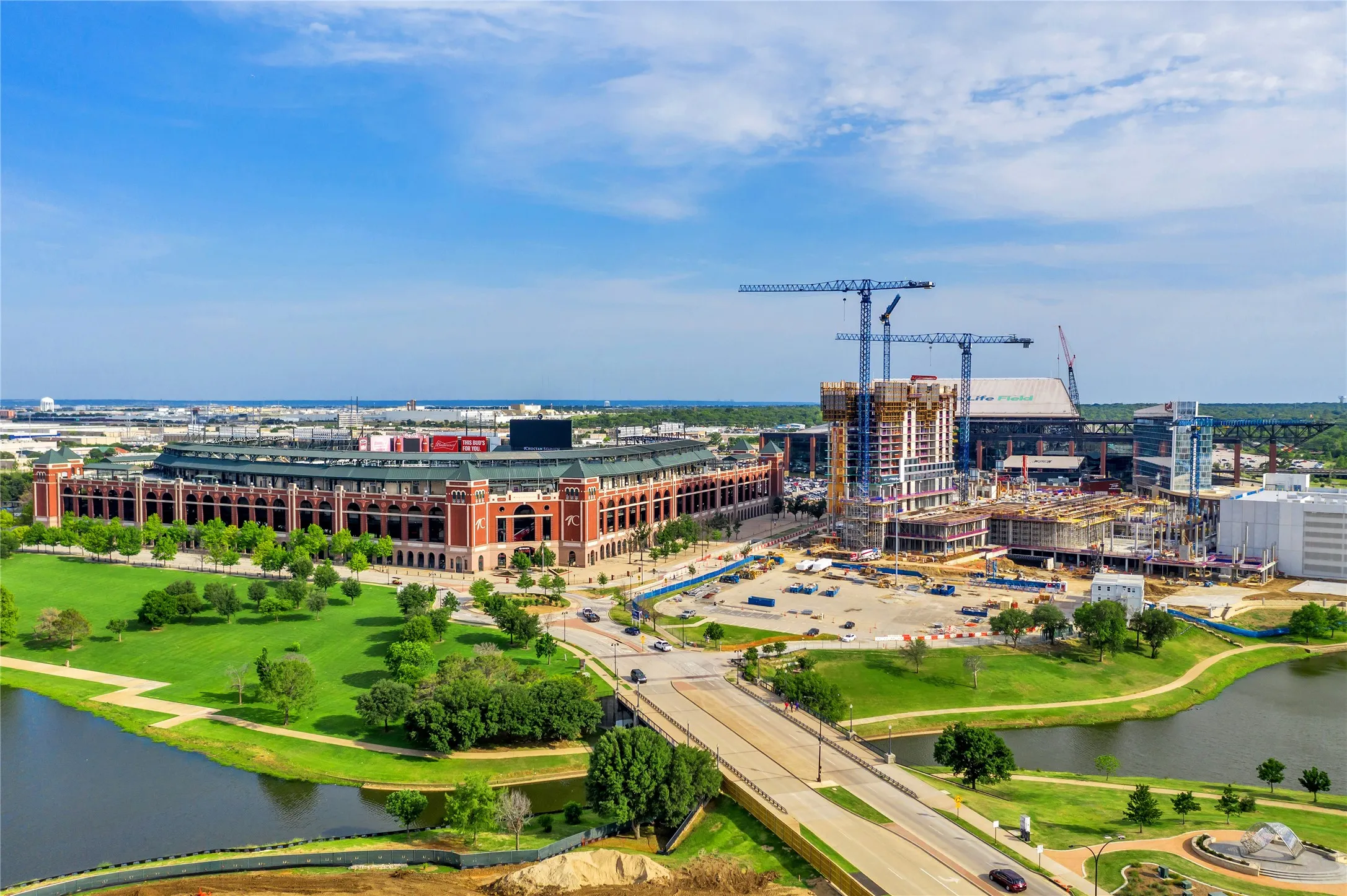 Aerial perspective showcasing a large stadium building, surrounding green spaces, a body of water, and active construction sites with multiple cranes