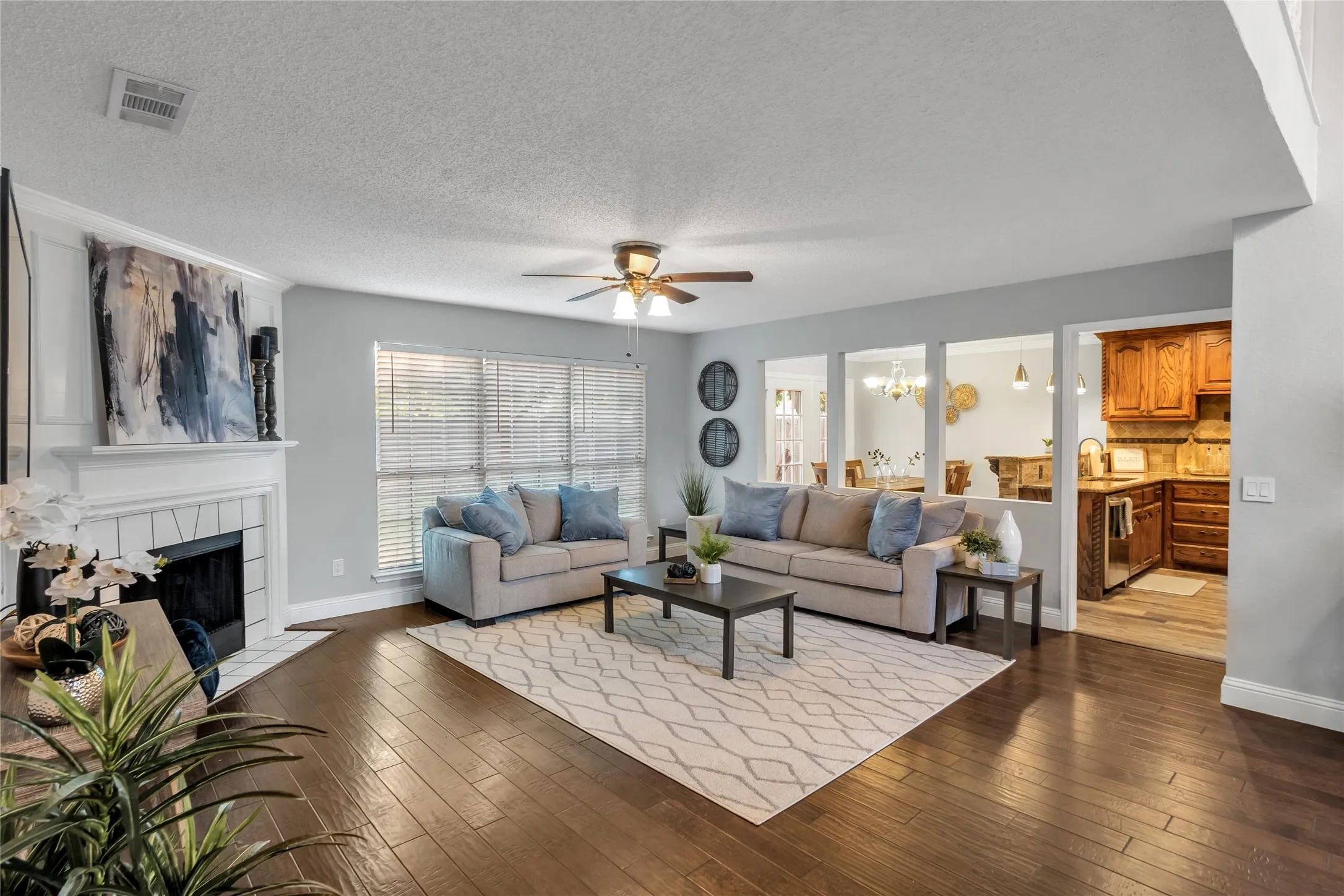 Spacious living area featuring wood-finish flooring, a white mantel fireplace, large windows with blinds, a ceiling fan, and open doorways to an adjacent kitchen with wood cabinetry