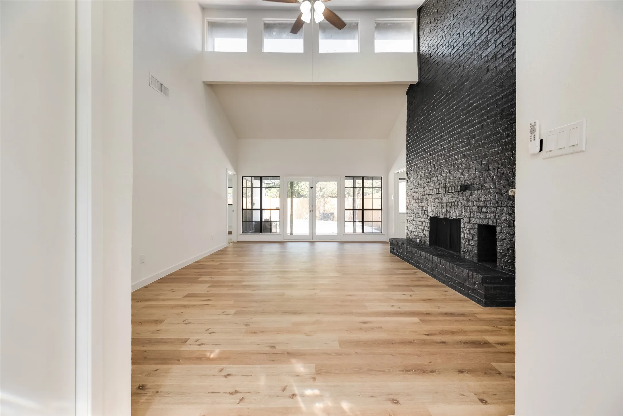 Vaulted ceiling with a ceiling fan, brick fireplace with a raised hearth, wood-finish flooring, and large windows with transom windows above