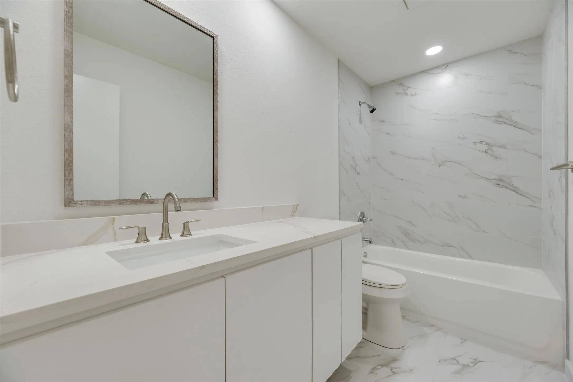 White vanity with integrated sink, brushed nickel faucet, large framed mirror, white bathtub with wall-mounted shower, and marble-look tile surround