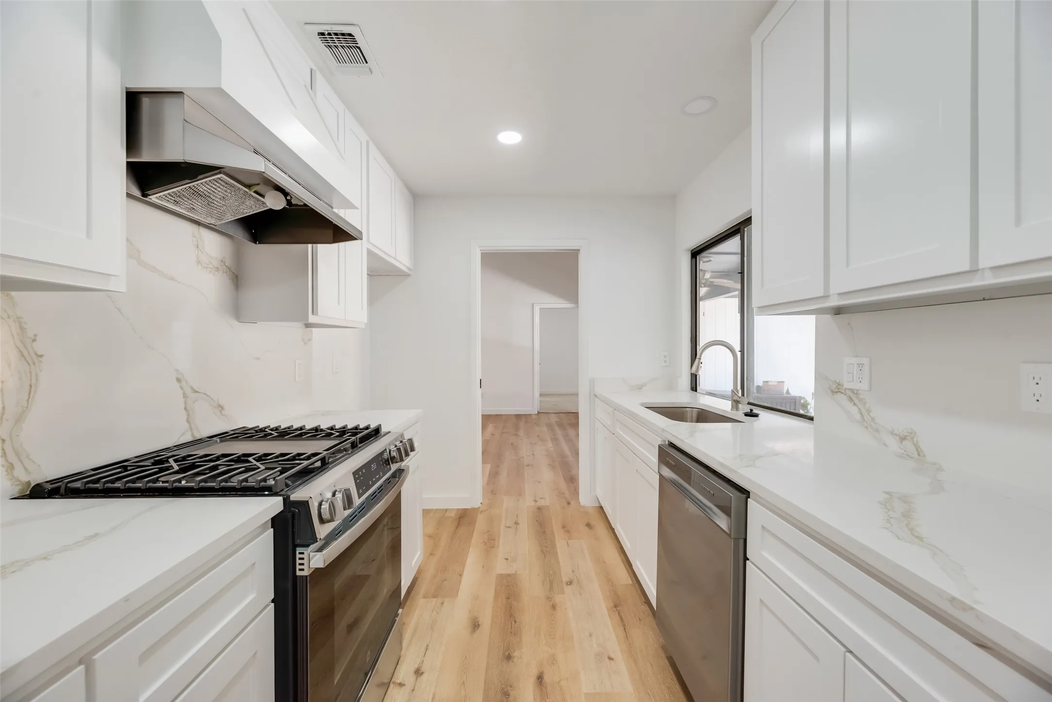 Galley kitchen featuring white cabinetry, quartz countertops, full-height backsplash, wood-finish flooring, and stainless steel appliances