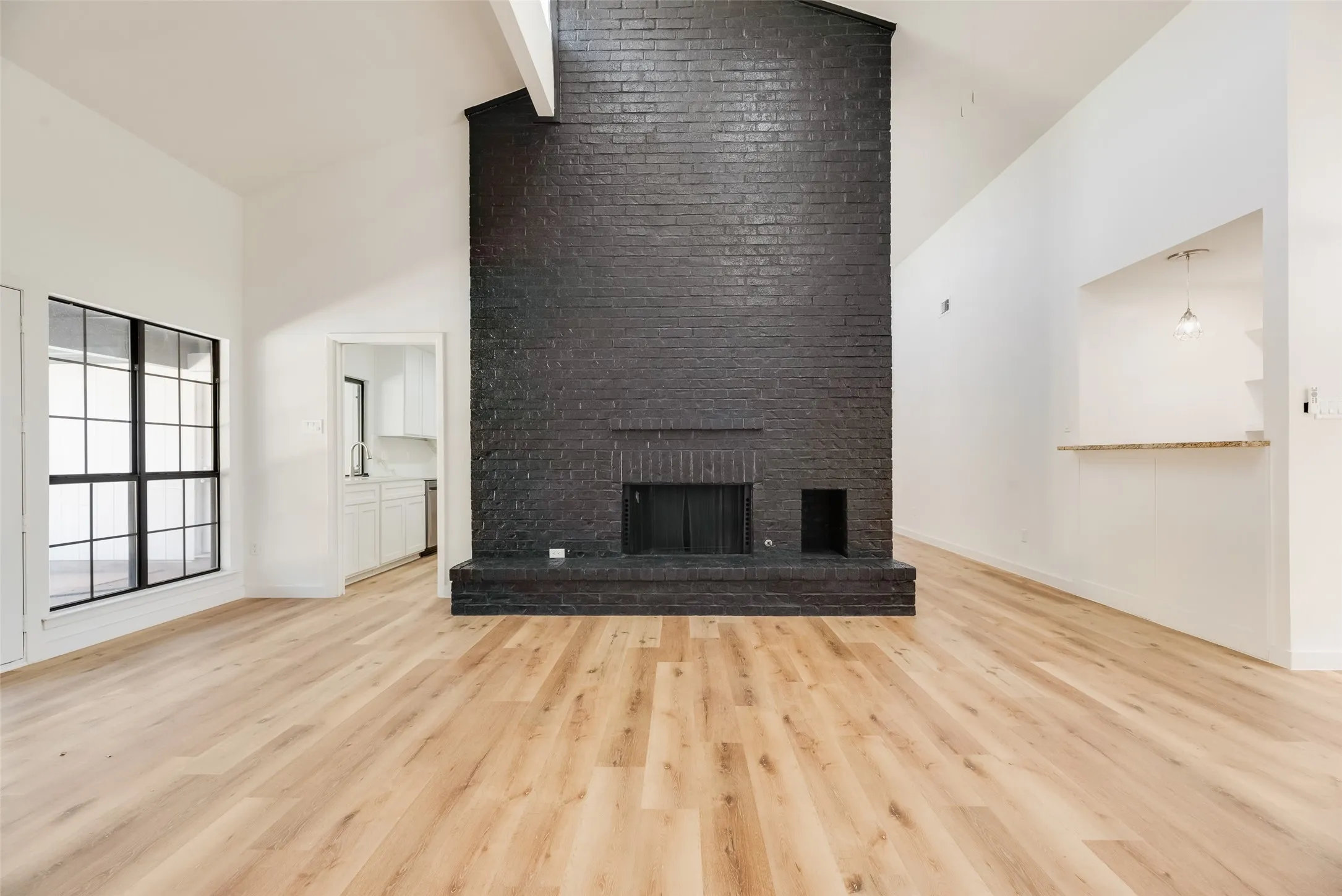 Vaulted ceilings, wood-finish flooring, a black painted brick fireplace, a pass-through window with a stone-finish countertop, and a large multi-pane window
