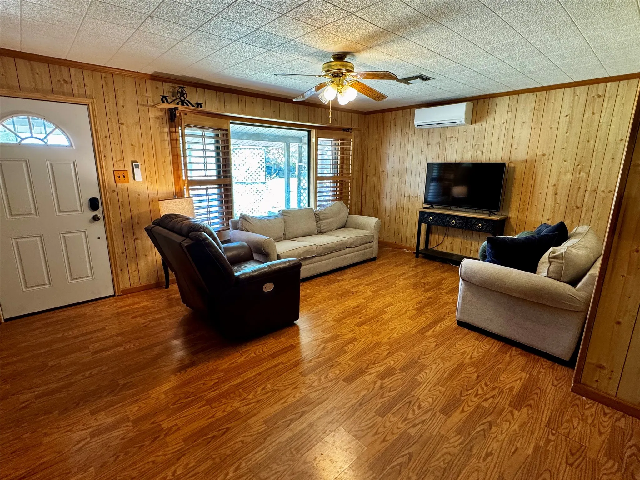 Wood-finish flooring, wood-paneled walls, a ceiling fan with integrated lighting, and a white paneled entry door with an arched window