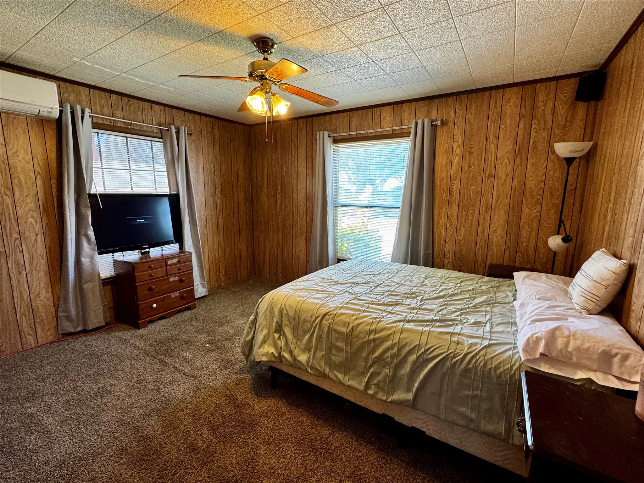 Wood paneling walls, textured ceiling, ceiling fan with integrated lighting, carpeted flooring, and a window with drapes