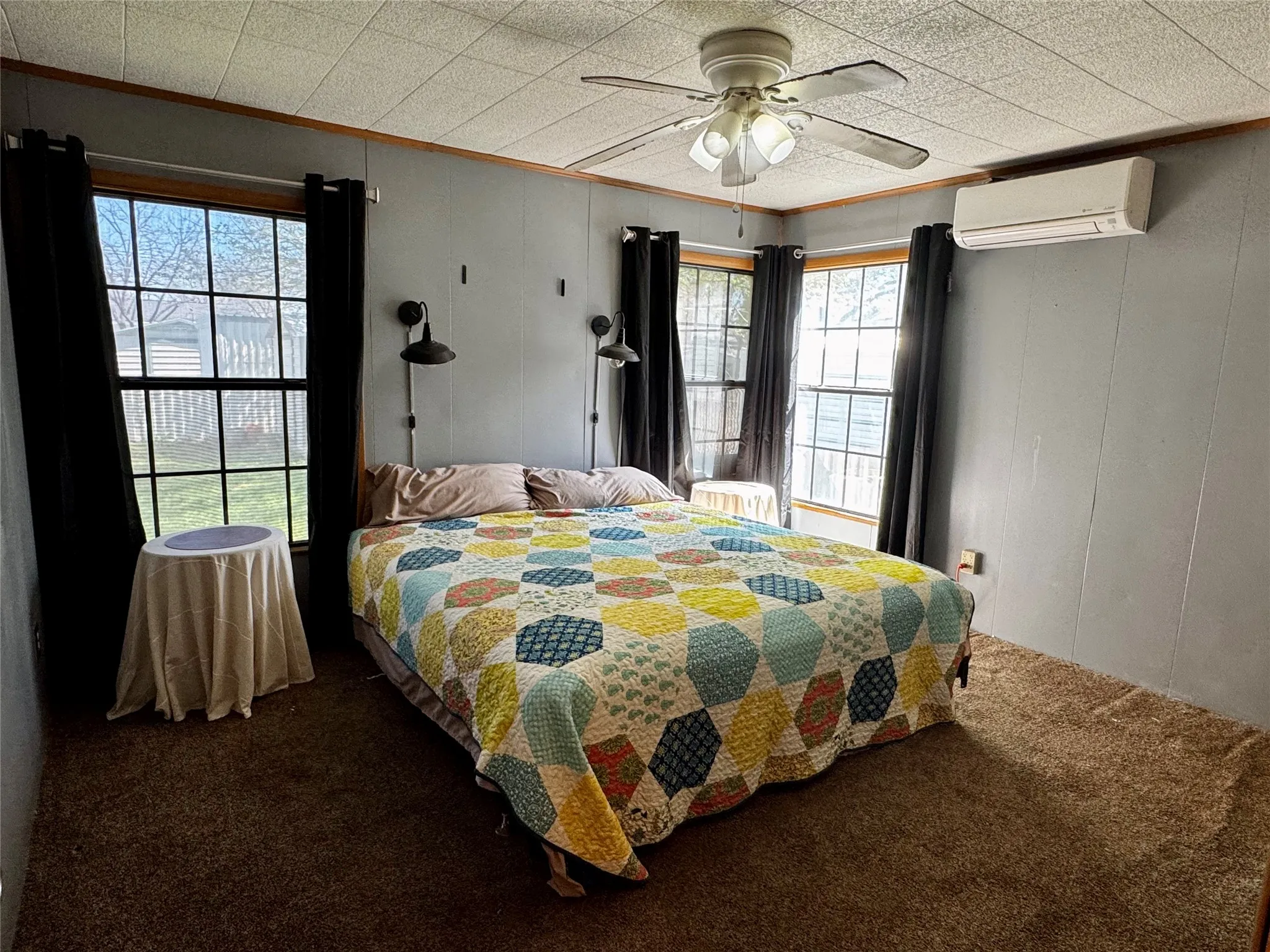 Ceiling fan with integrated lighting, wall-mounted mini-split system, two windows, textured wall panels, and carpeted flooring