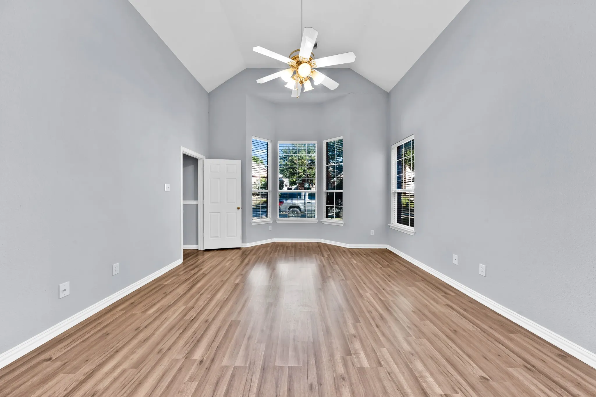 Vaulted ceiling, wood-finish flooring, bay window, white trim, and a ceiling fan with integrated lighting