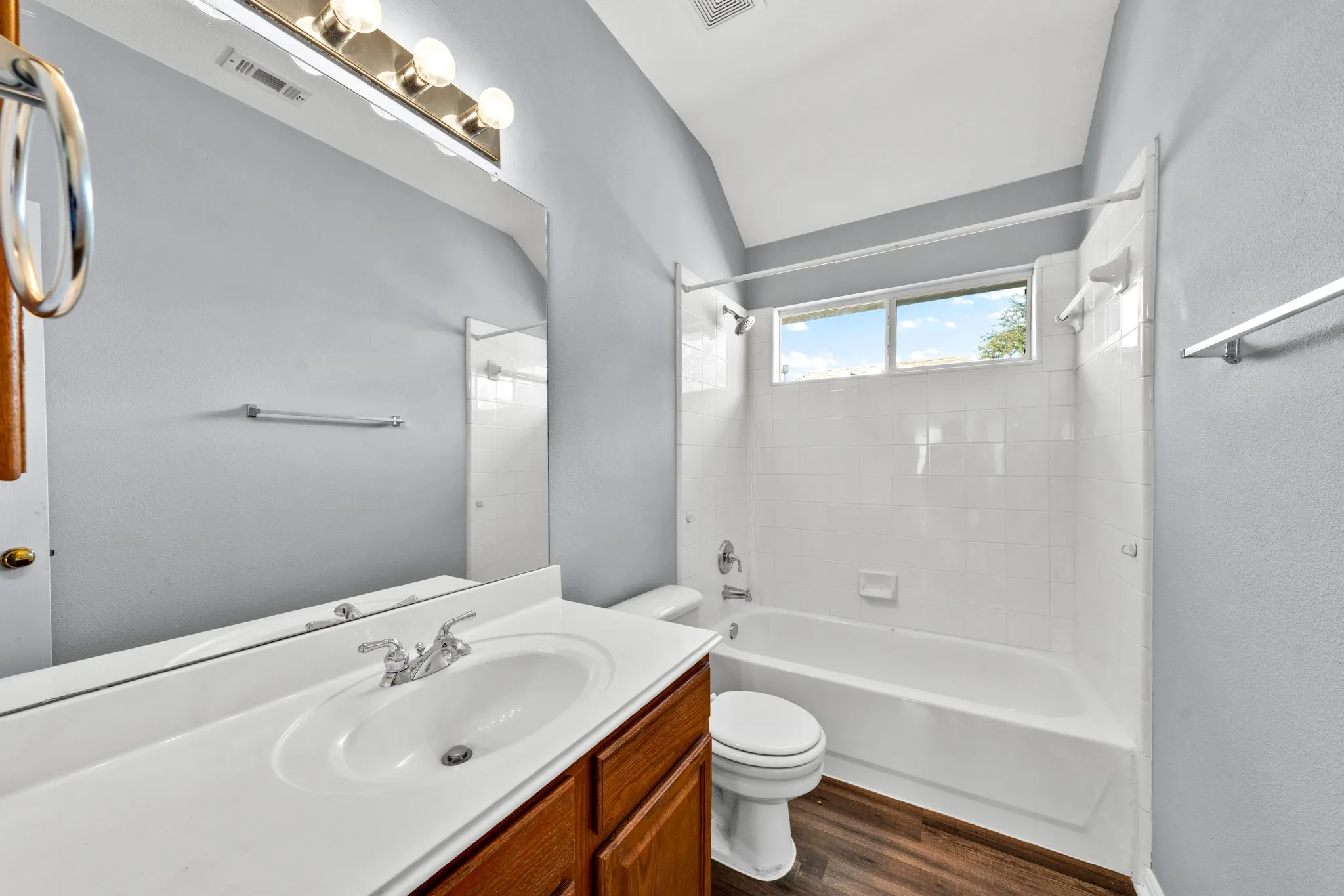 Bathroom featuring a single vanity with wood-finish cabinetry, a large frameless wall mirror, a bathtub with a shower, and wood-finish flooring