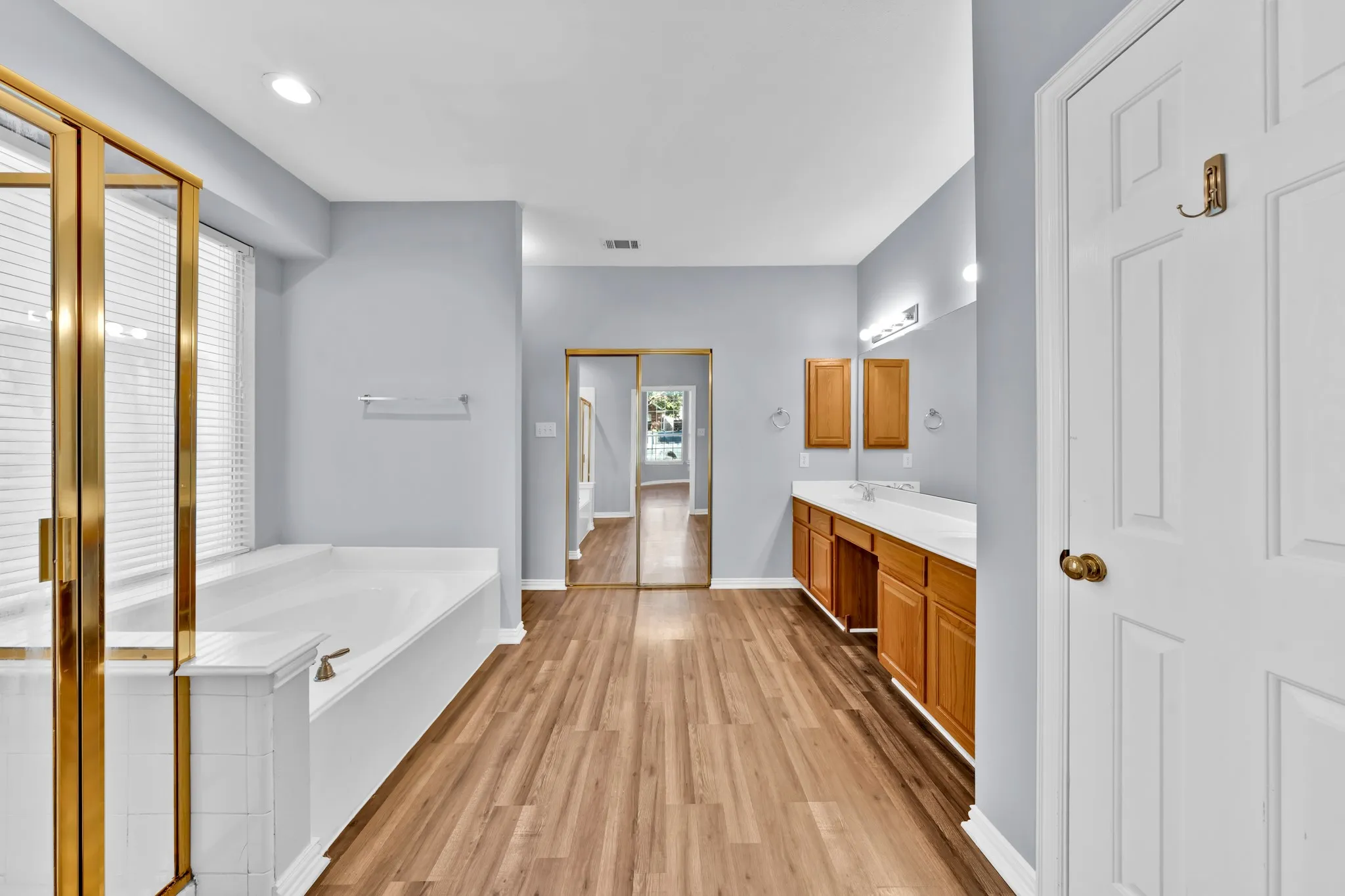 Integrated soaking tub with tiled surround, wood-finish flooring, dual vanity with wood cabinetry, full-height mirrored closet doors, and recessed lighting
