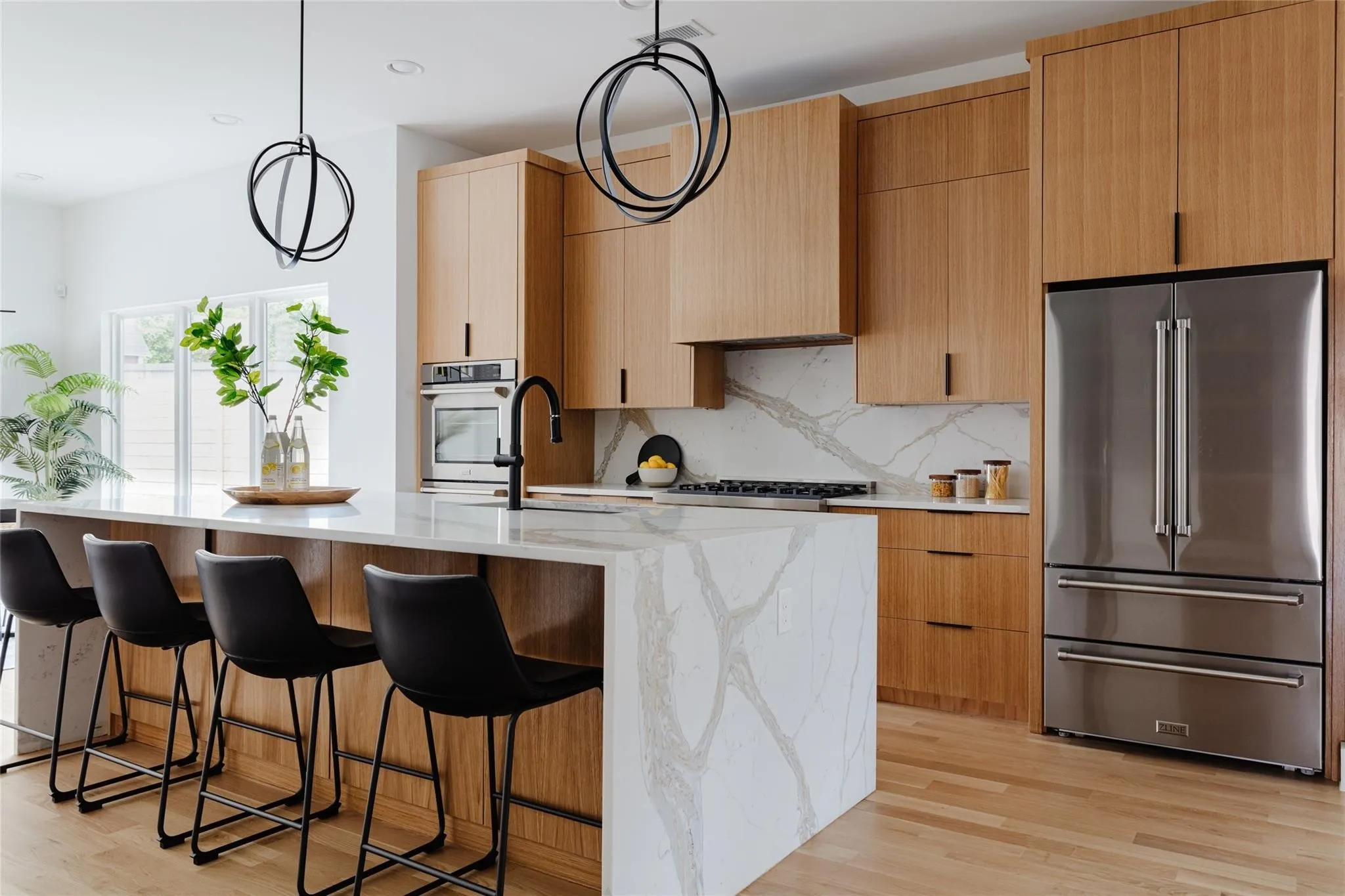 Light wood-finish cabinetry and flooring, stainless steel appliances, and a waterfall island with a patterned countertop