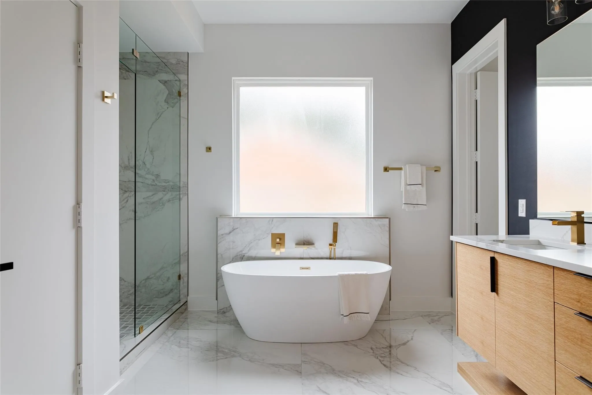 Freestanding soaking tub with wall-mounted brass fixtures, glass-enclosed shower with marble-look tile, double vanity with light wood-finish cabinetry, white countertop, and brass faucets, and marble-look floor tile throughout