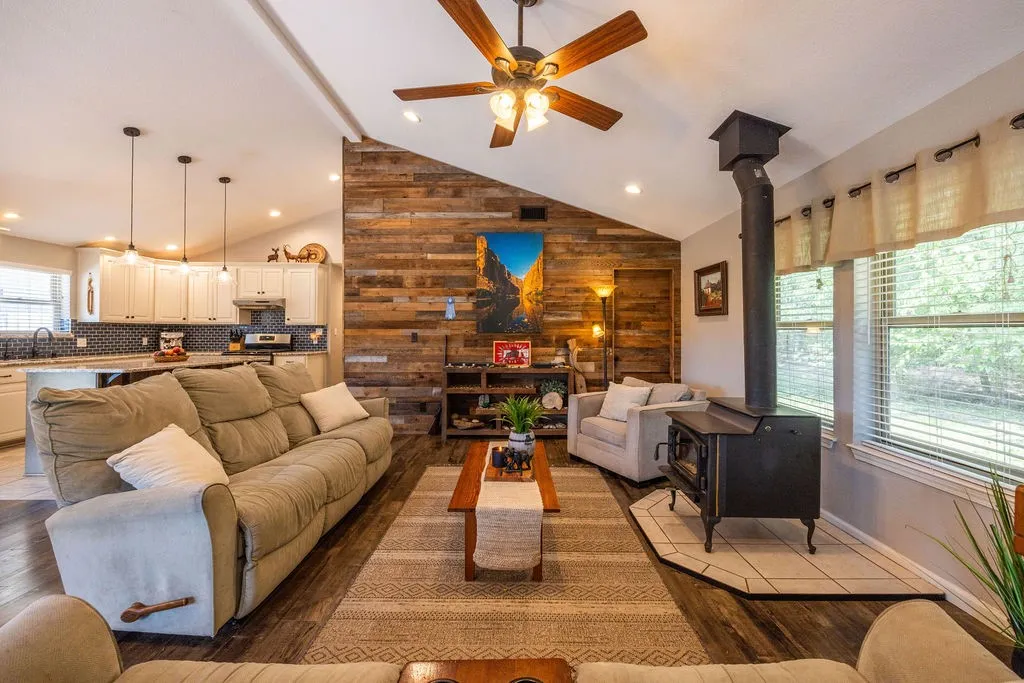 Vaulted ceiling with a ceiling fan, wood-finish flooring, wood-paneled accent wall, and a black wood-burning stove with a tile hearth
