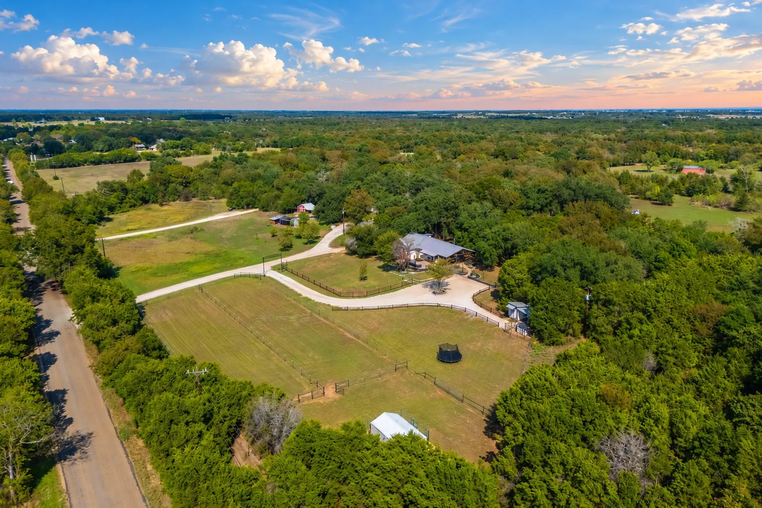 Property featuring extensive tree coverage, open fields, a main residence with an attached garage, and a long gravel driveway