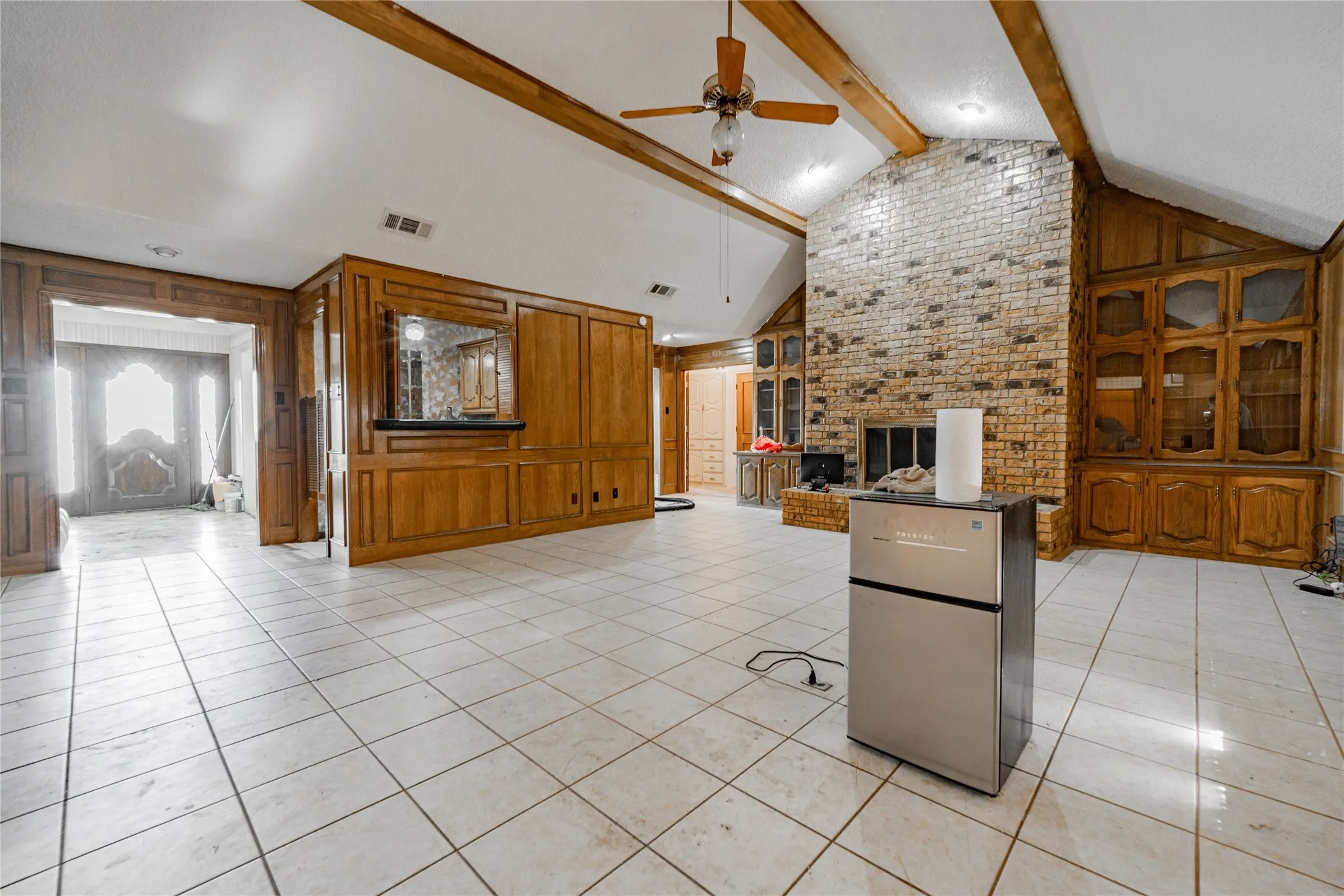 Vaulted ceiling with exposed wood beams, brick fireplace, built-in wood cabinetry, wood paneling, and tile flooring
