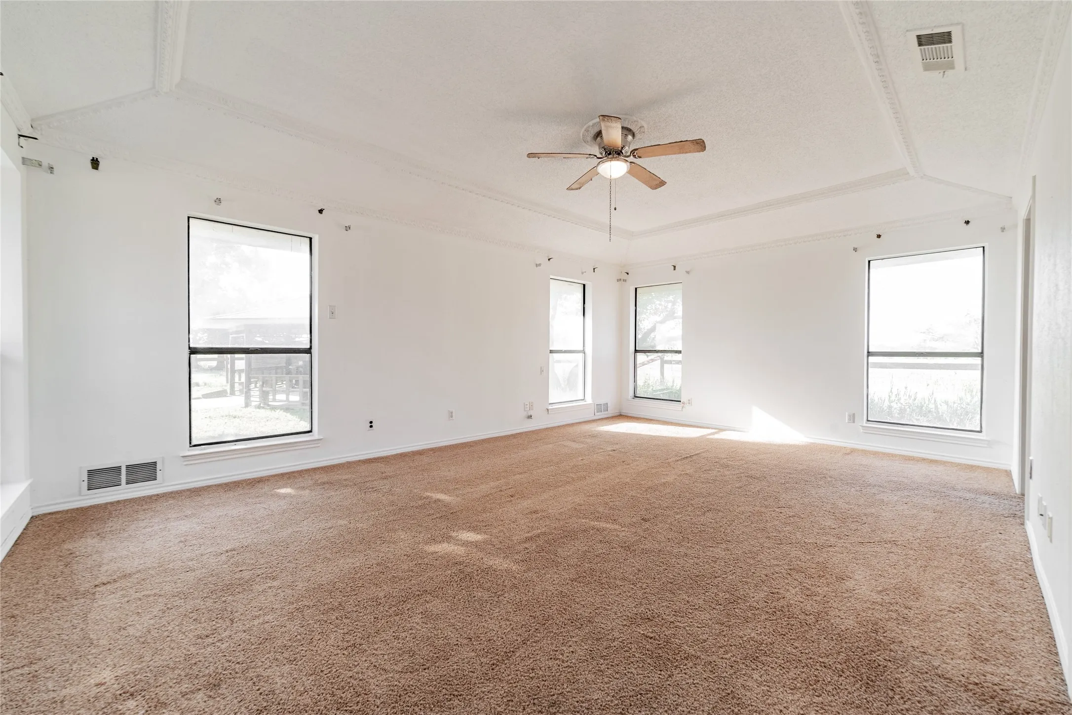 Spacious room featuring tray ceilings, neutral carpeting, a ceiling fan with integrated lighting, and multiple windows