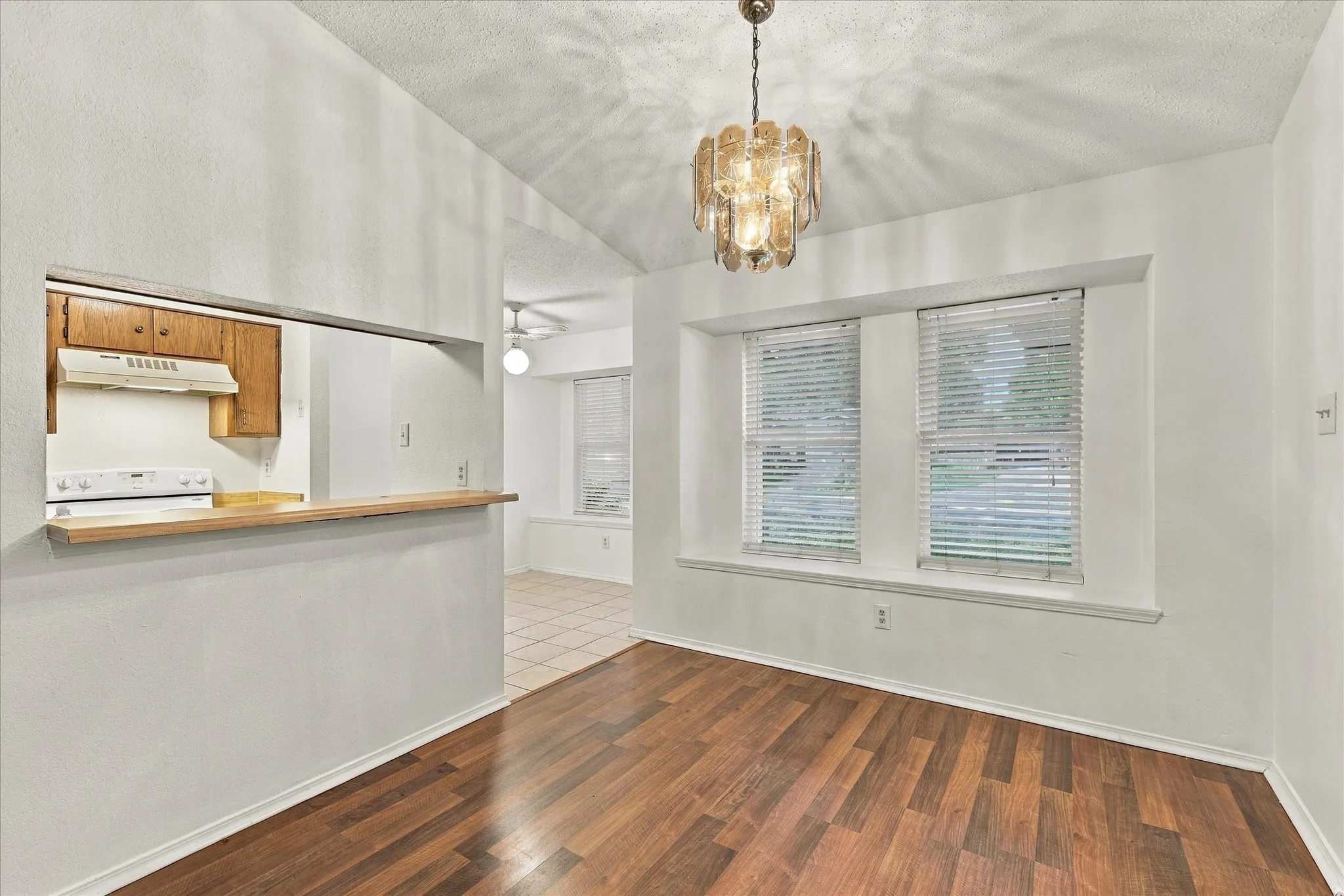 Vaulted ceiling with an ornate chandelier, wood-finish flooring, two large windows with blinds, and a pass-through opening to the kitchen area with wood cabinetry and a white range