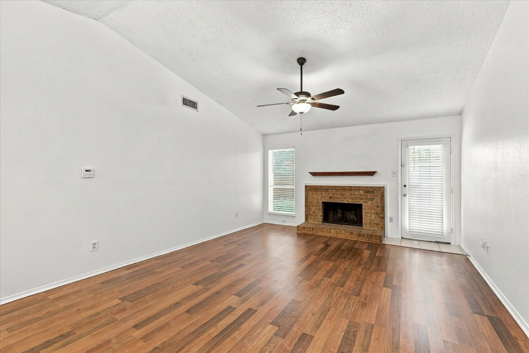Vaulted ceiling with a ceiling fan, brick fireplace with a wood mantel, wood-finish flooring, and multiple windows