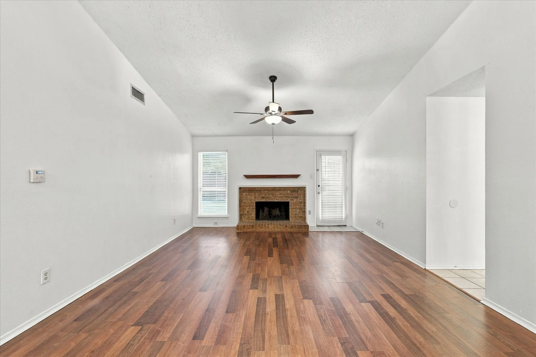 Vaulted ceiling, wood-finish flooring, brick fireplace with wood mantel, ceiling fan, and large window