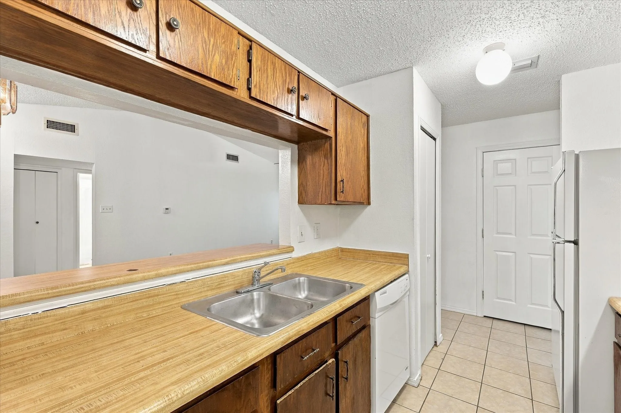 Kitchen featuring wood cabinetry, a pass-through window, a double basin stainless steel sink, tiled flooring, and a white refrigerator