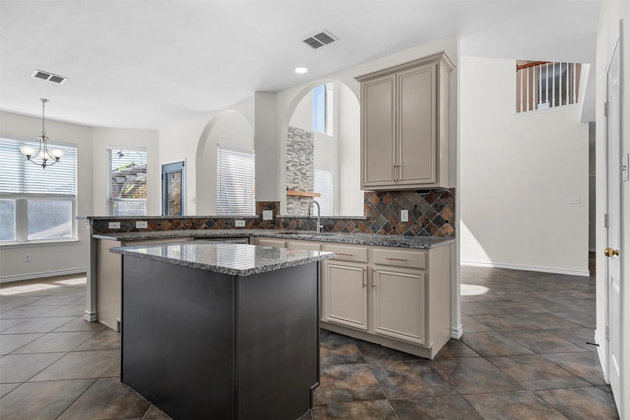 Expansive kitchen featuring a central island with a stone-finish countertop, coordinating perimeter countertops, light cabinetry, and a mosaic tile backsplash