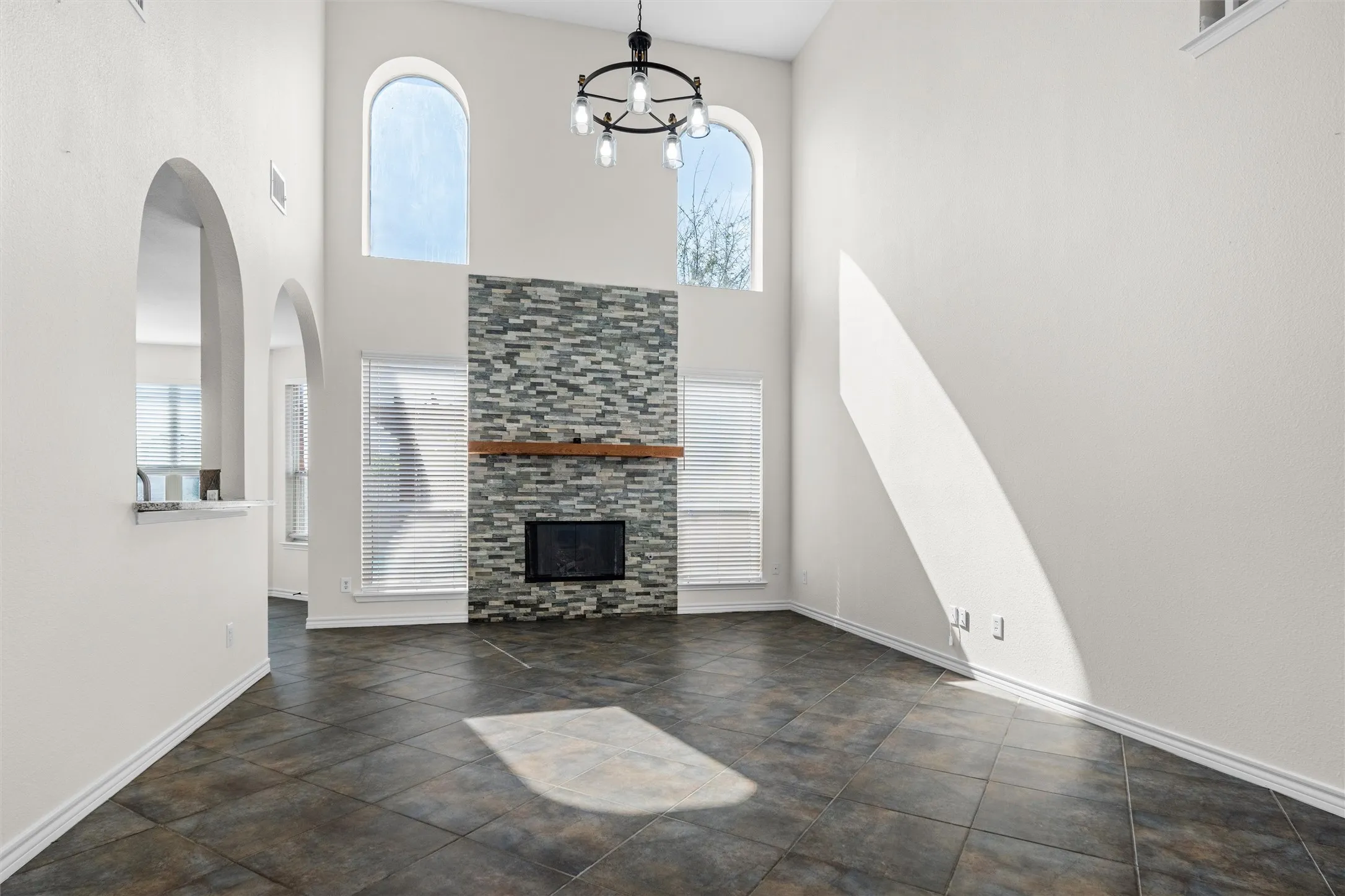 Vaulted ceiling with a contemporary chandelier, arched windows, tile flooring, and a fireplace with a stone surround and wood mantle