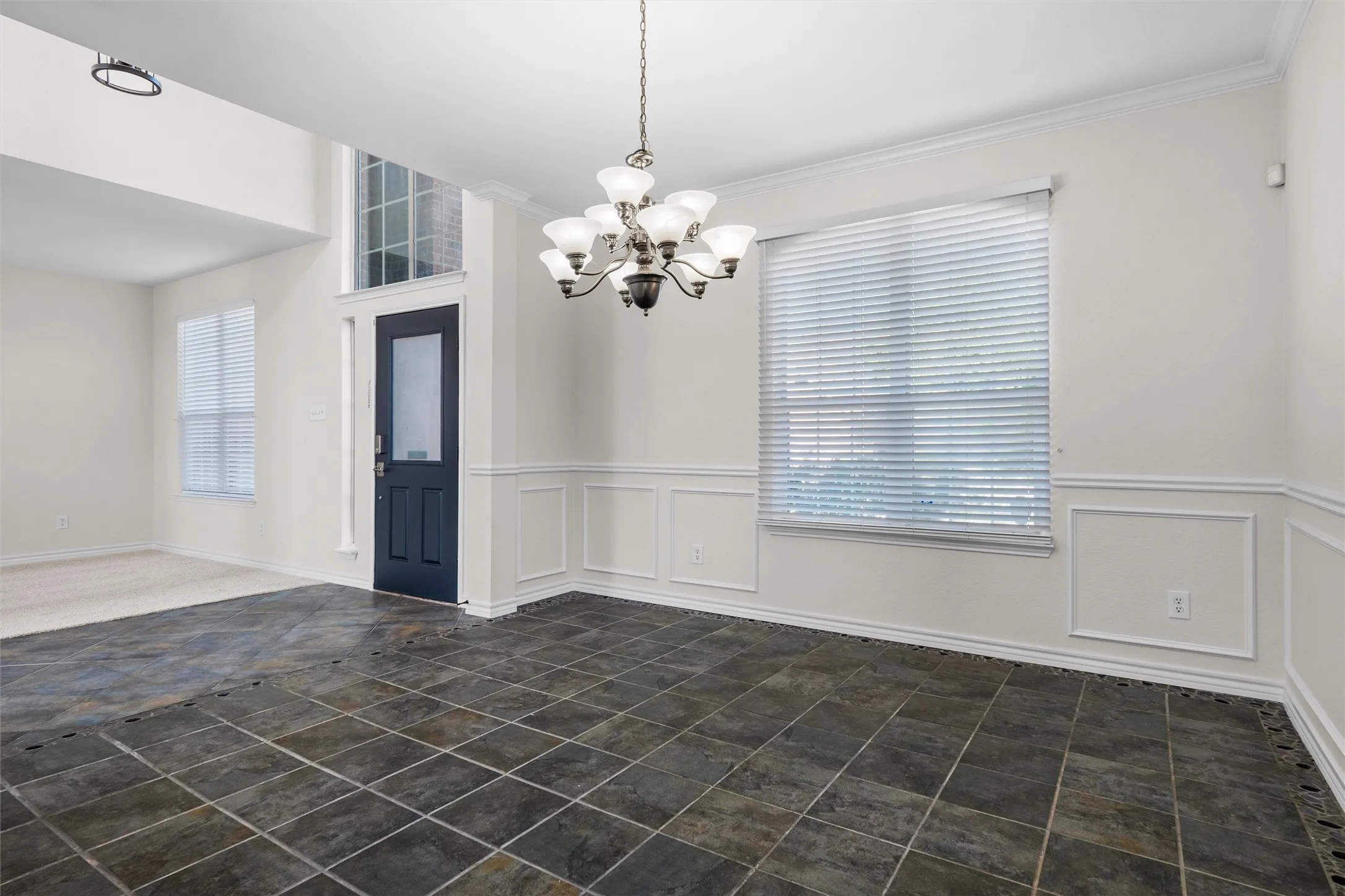 Tile flooring, decorative wall paneling, window with blinds, front door with glass insert, and a chandelier
