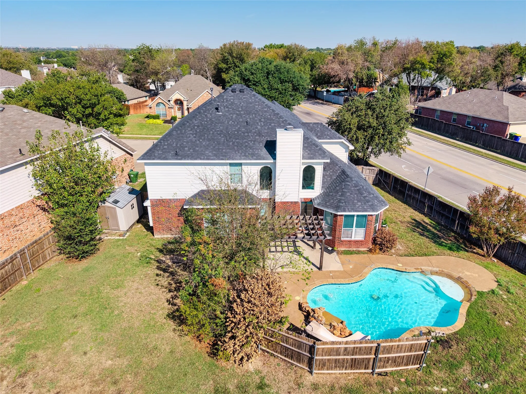 Brick and siding exterior with a dark shingle roof, featuring a built-in outdoor pool, wood pergola, and a storage shed