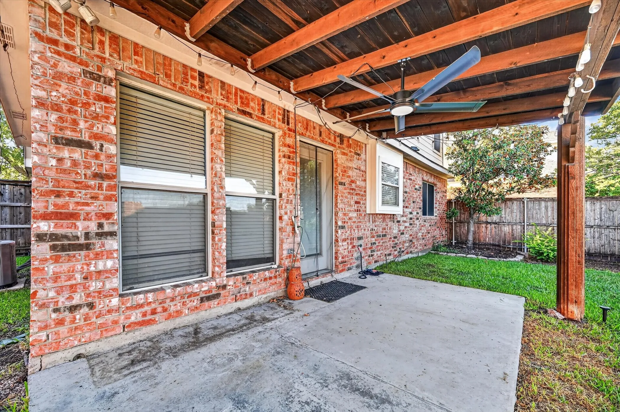 Expansive concrete patio with a wood-beamed overhead structure, integrated ceiling fan, and brick exterior