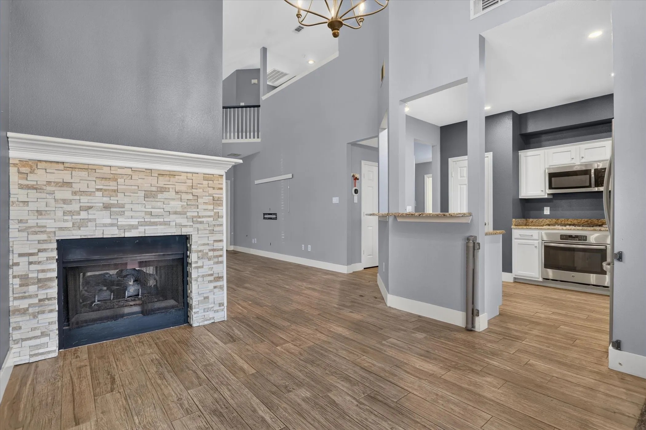 Floor-to-ceiling fireplace with stacked-stone facade, wood-finish flooring, soaring ceilings, kitchen featuring white cabinetry and stone countertops, and overhead decorative lighting