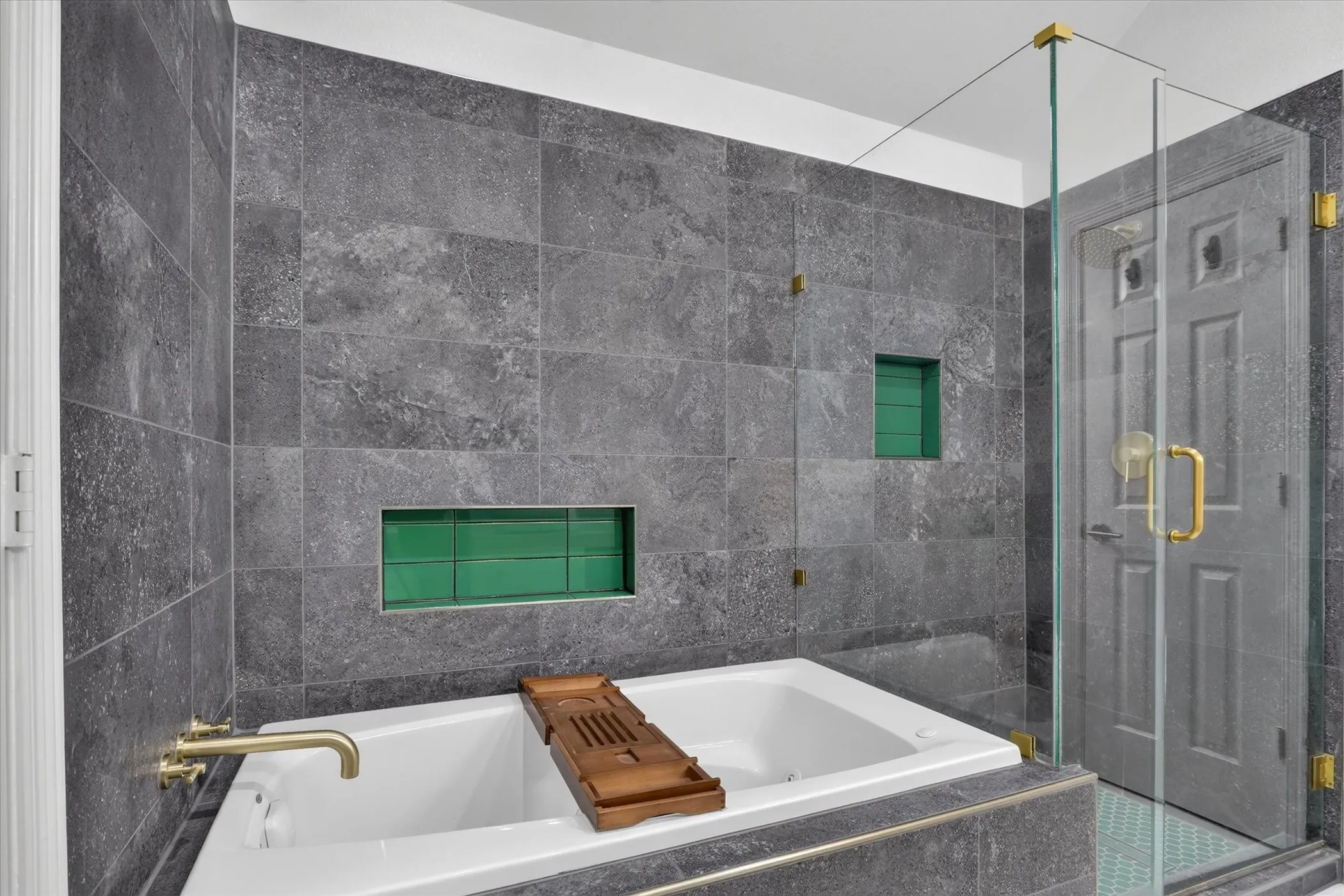 Large soaking tub featuring gold-tone hardware, dark stone-finish wall tiling, glass-enclosed shower with gold-tone fixtures, and dual recessed wall niches