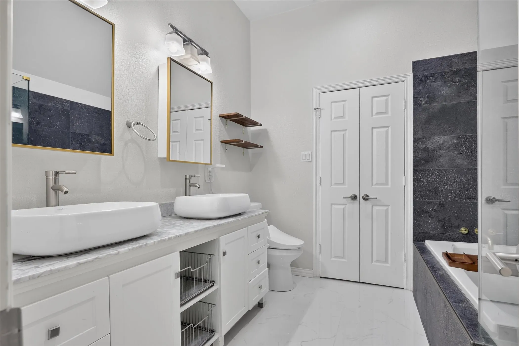 Dual vanity with rectangular vessel sinks, stone-finish countertop, white cabinetry with built-in wire storage, wall-mounted light fixtures, and a soaking tub featuring dark-toned stone-finish tile surround