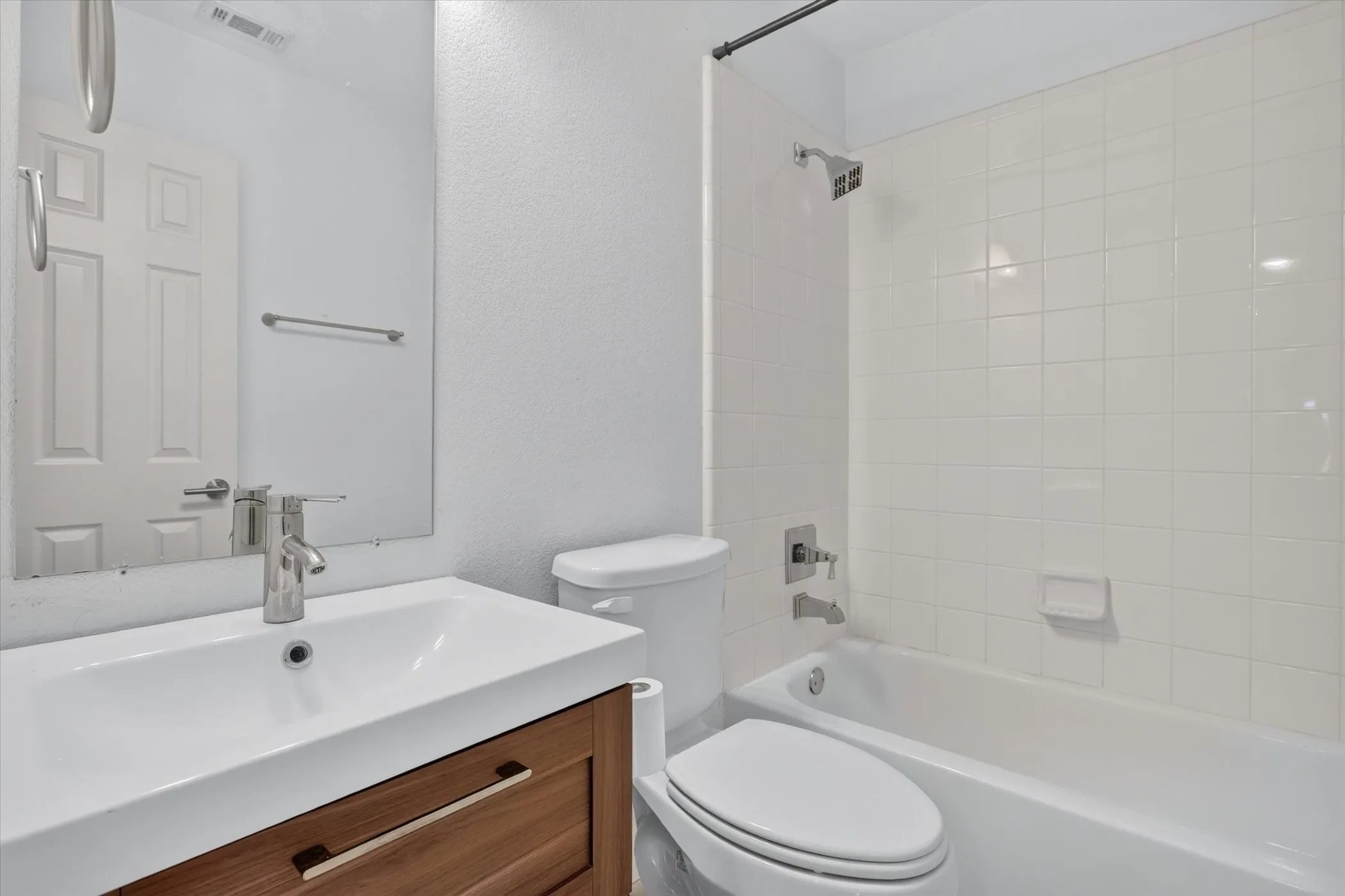 Bathroom featuring a wood-finish floating vanity, rectangular sink with a chrome faucet, large frameless wall-mounted mirror, and a tub-shower combination with white tiled surround