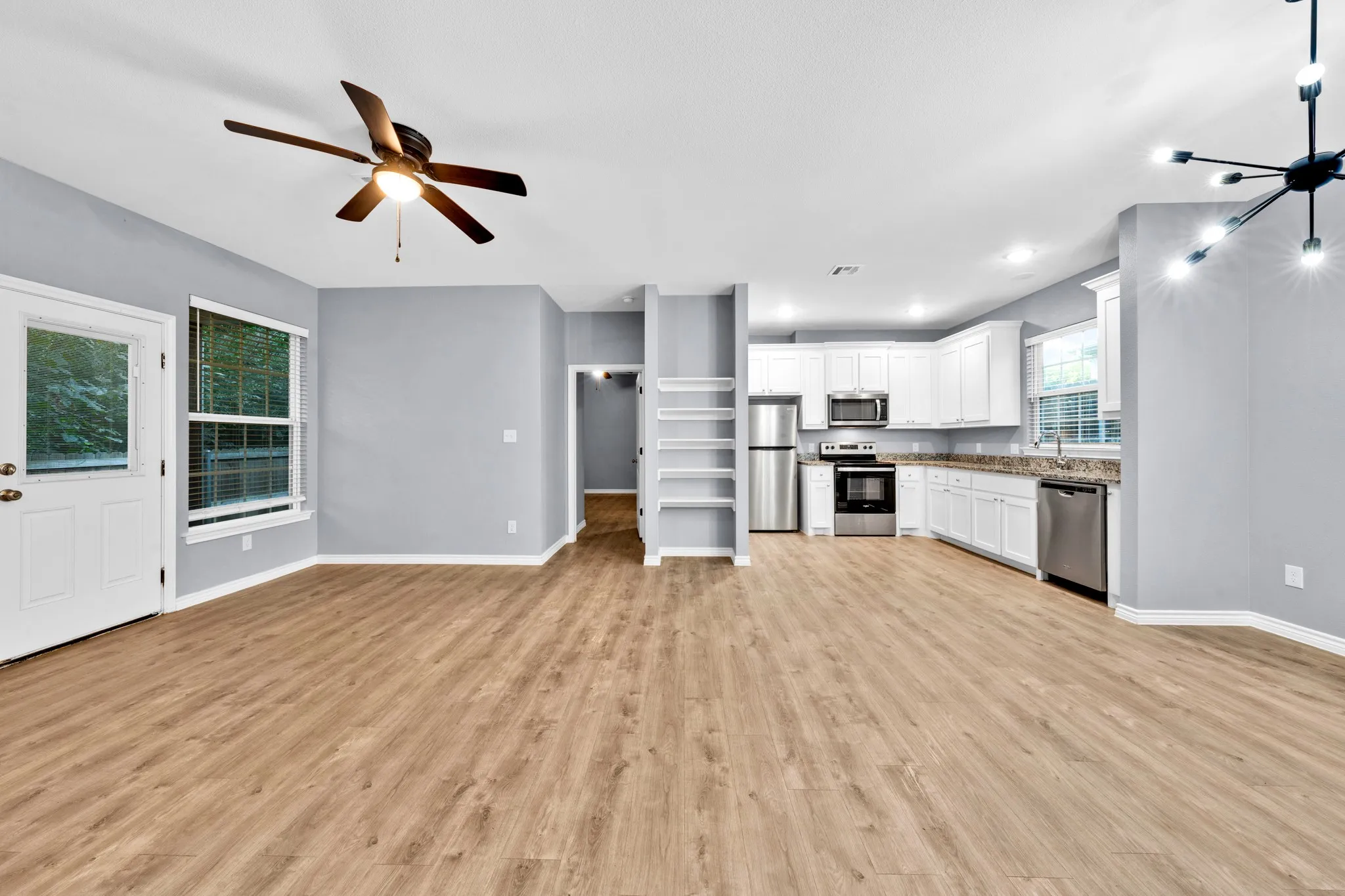 Open-concept living space with wood-finish flooring, a ceiling fan, a contemporary chandelier, white cabinetry, and stainless steel appliances