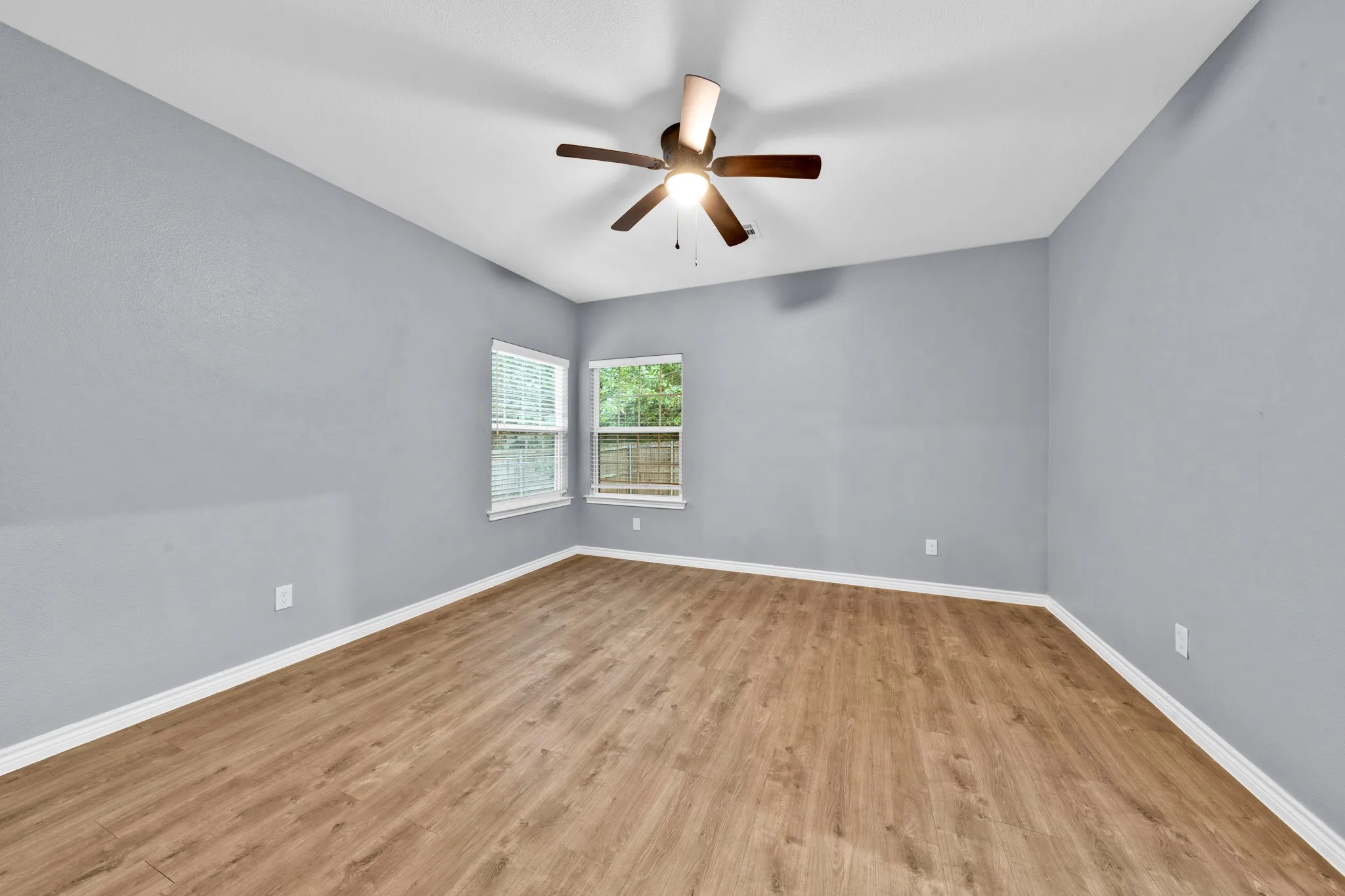 Wood-finish flooring, two windows with blinds, ceiling fan with integrated lighting, and light gray walls