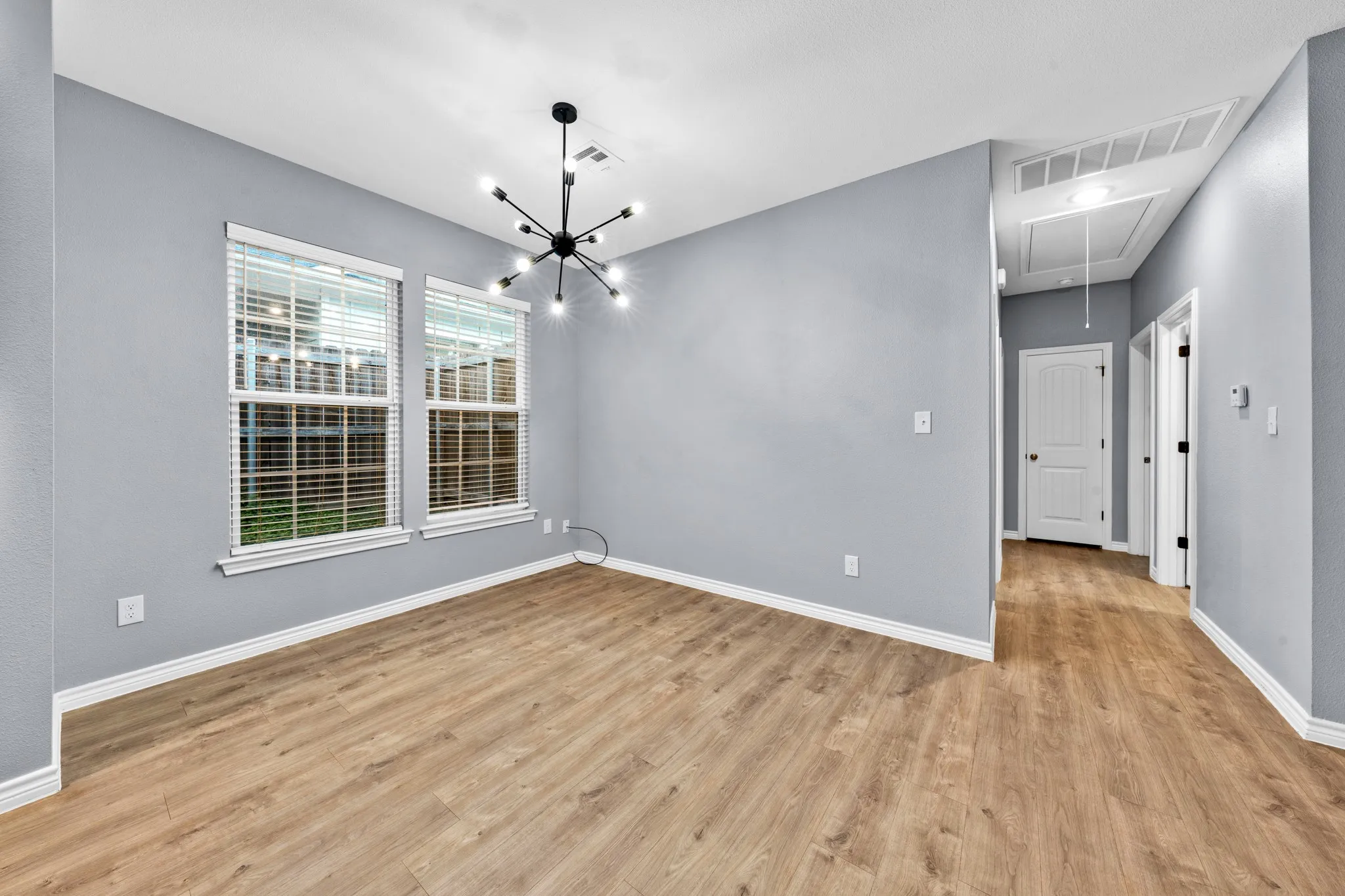 Spacious room featuring wood-finish flooring, two large windows with blinds, a modern sputnik chandelier, light gray wall paint, and white trim