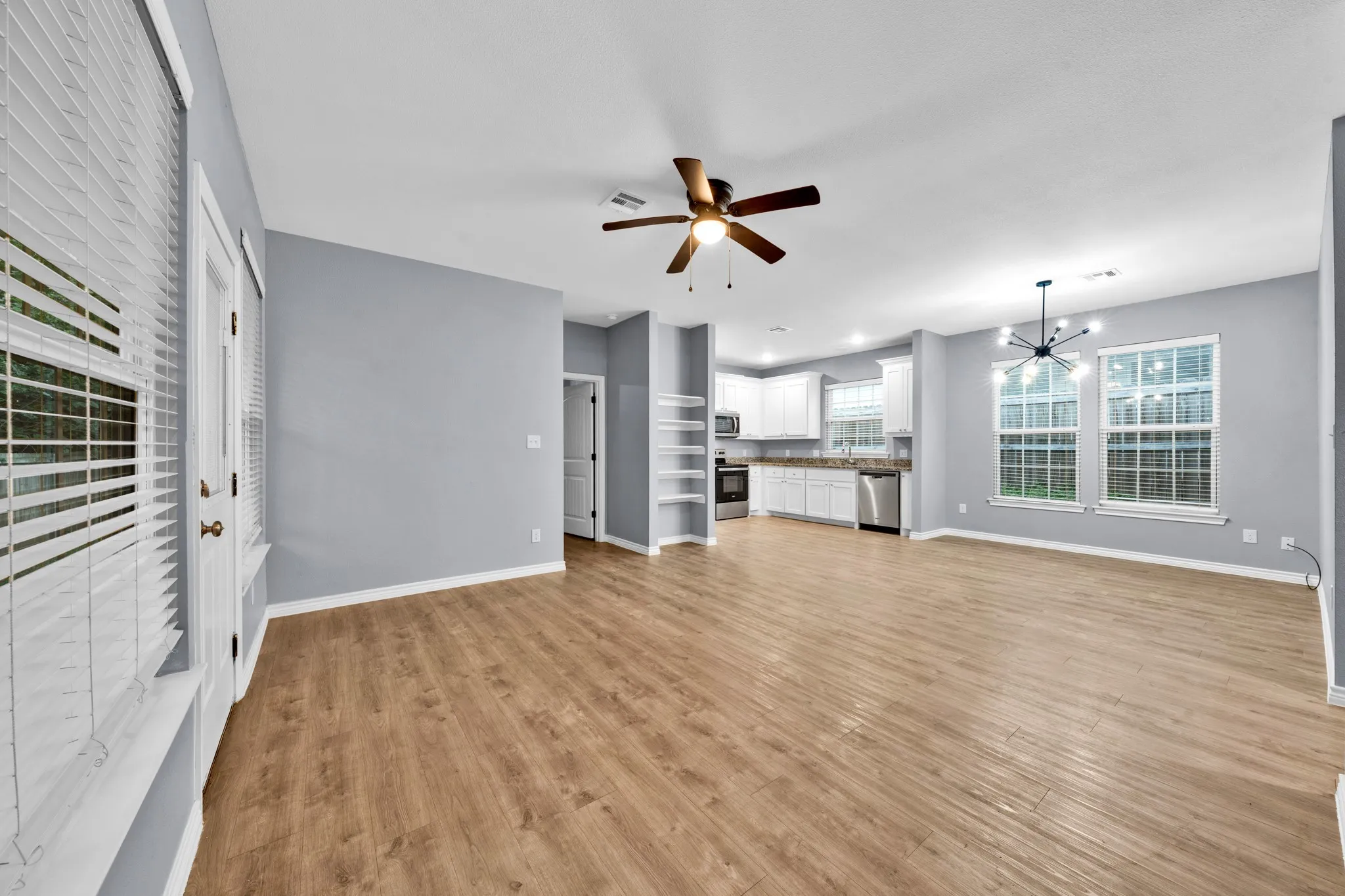 Open-concept living space with wood-finish flooring, white cabinetry, stainless steel appliances, and an exposed bulb chandelier