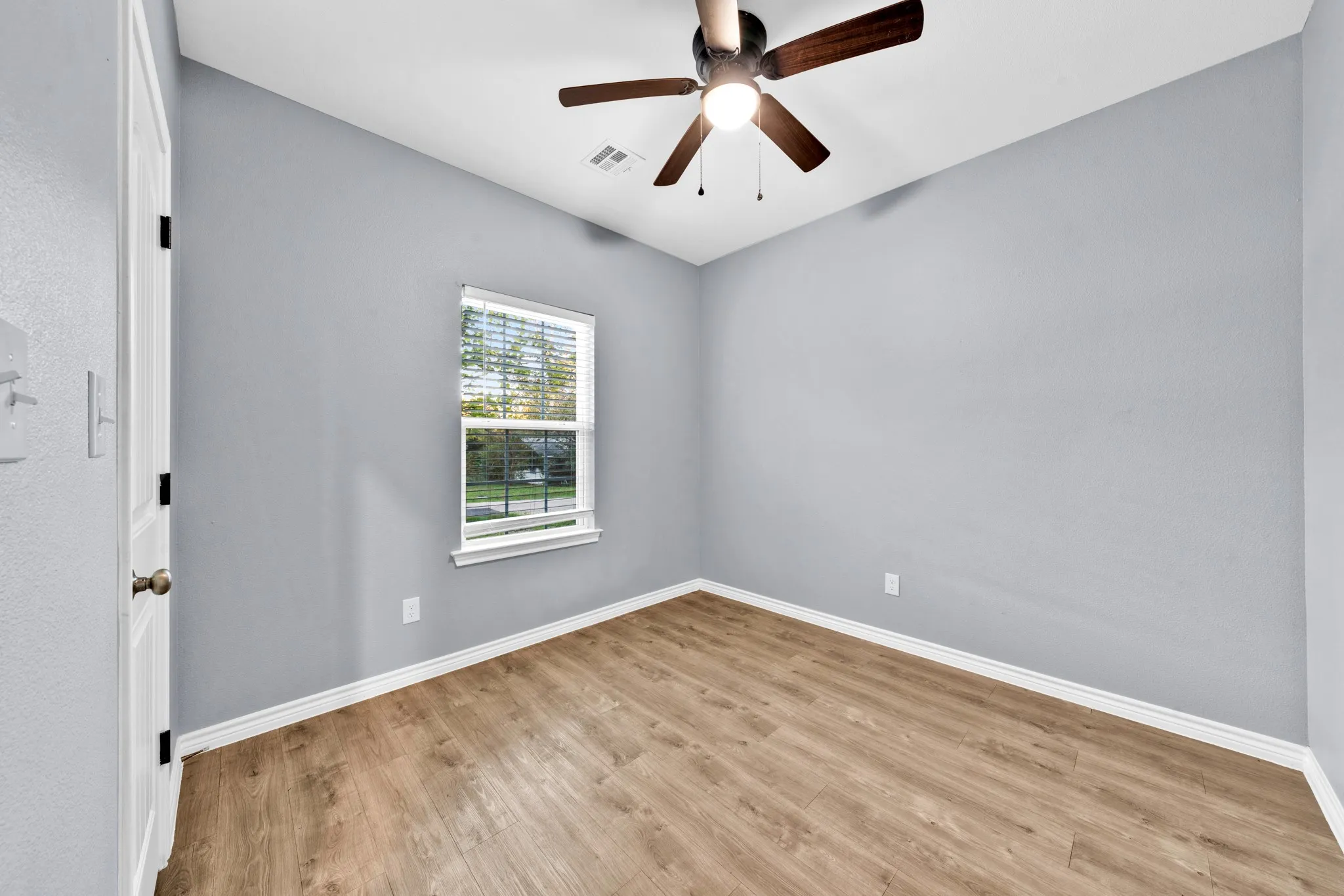 Wood-finish flooring, light gray wall paint, window with blinds, and a ceiling fan with light fixture