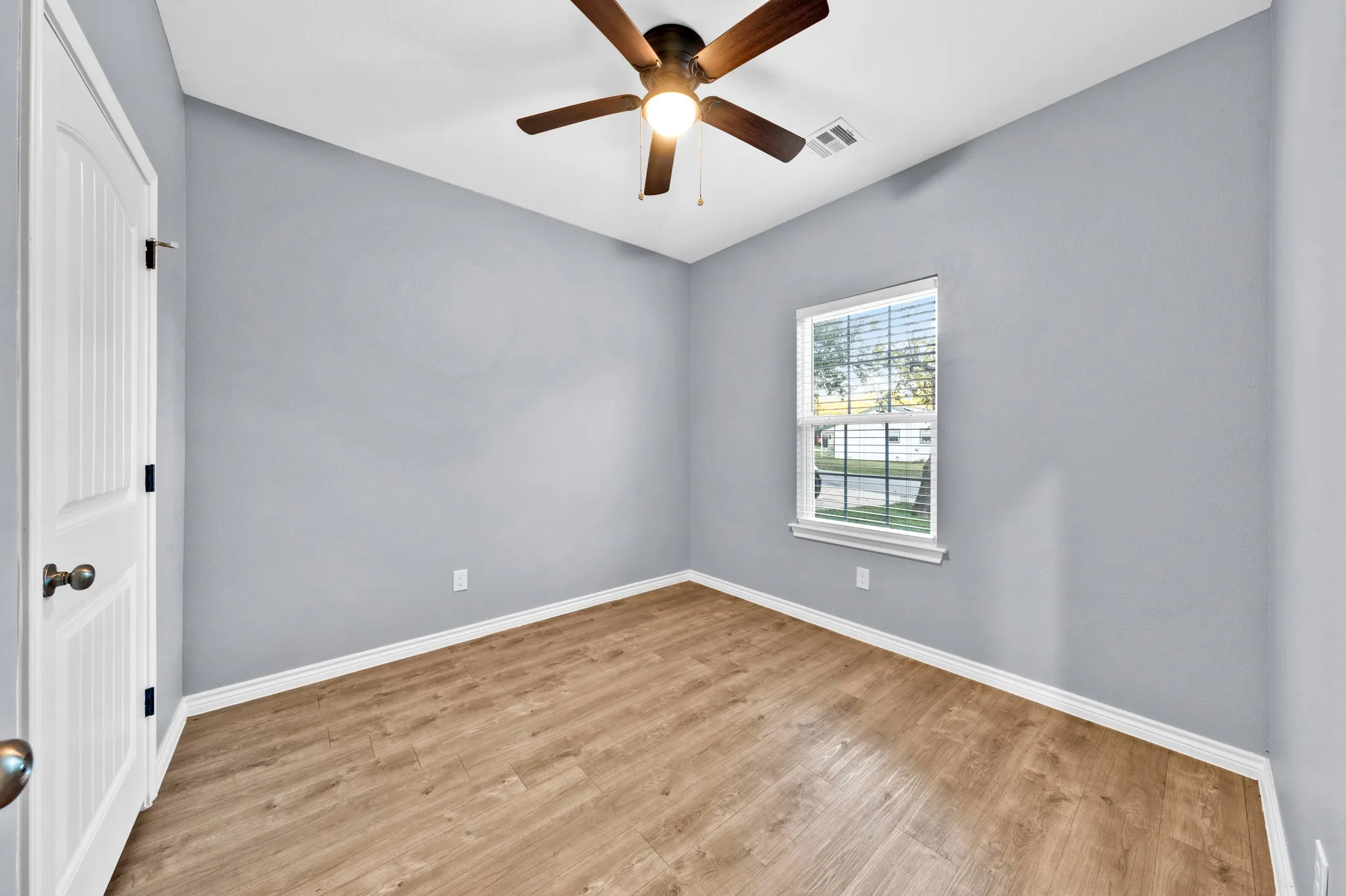 Wood-finish flooring, single window with blinds, ceiling fan with integrated lighting, paneled door with dark hardware