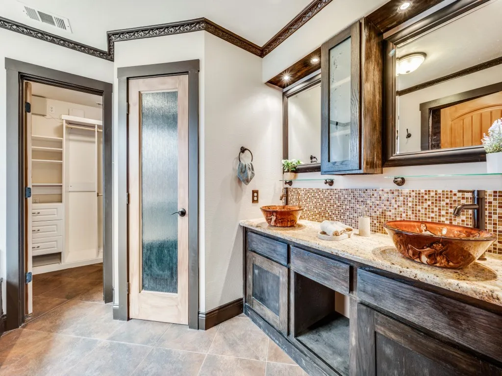 Dual vessel sinks, tile flooring, mosaic tile backsplash, custom wood cabinetry, and a walk-in closet with built-in shelving