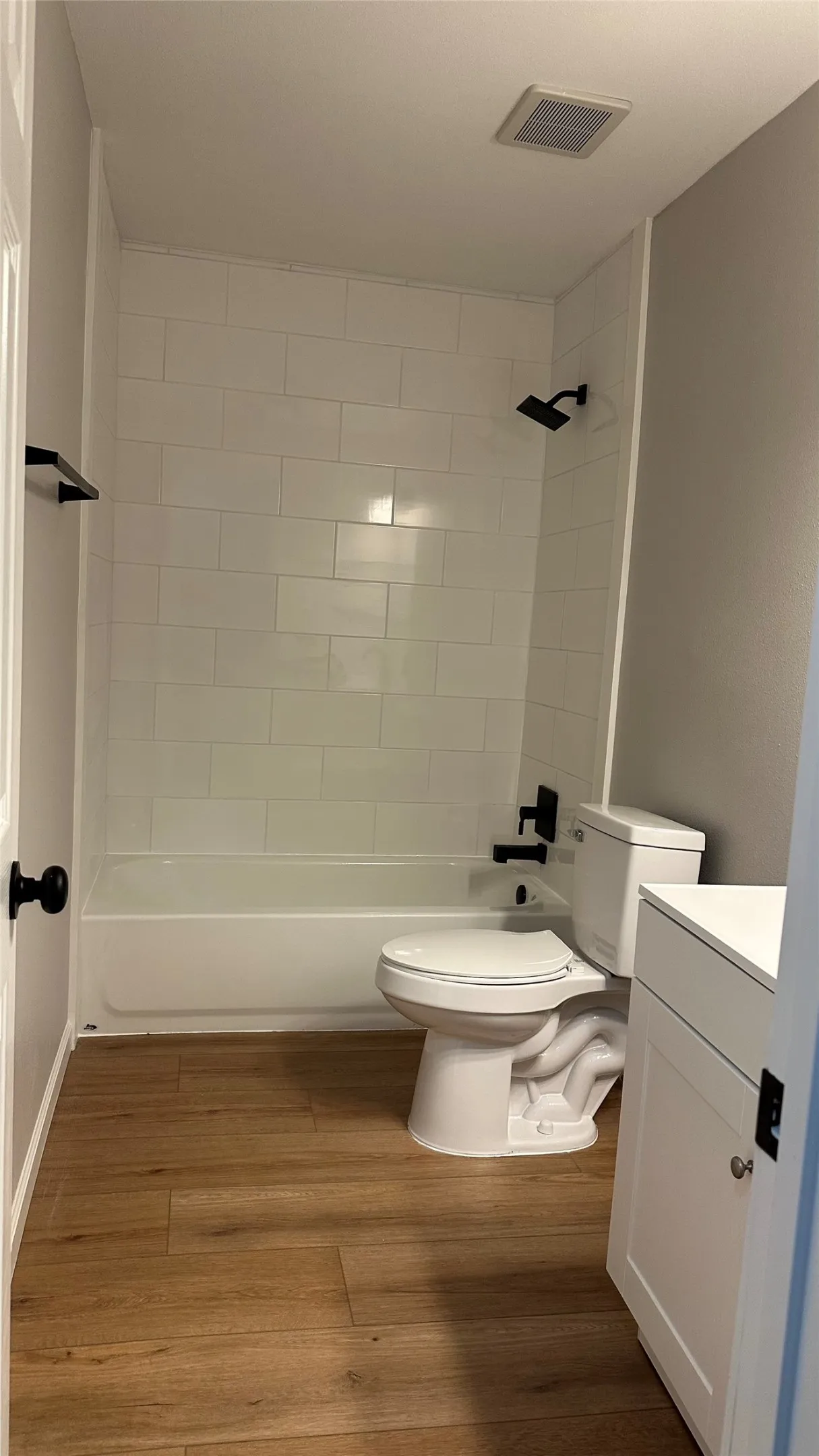 Wood-finish flooring, white subway tile shower surround, white bathtub, white vanity with shaker-style door, and matte black fixtures