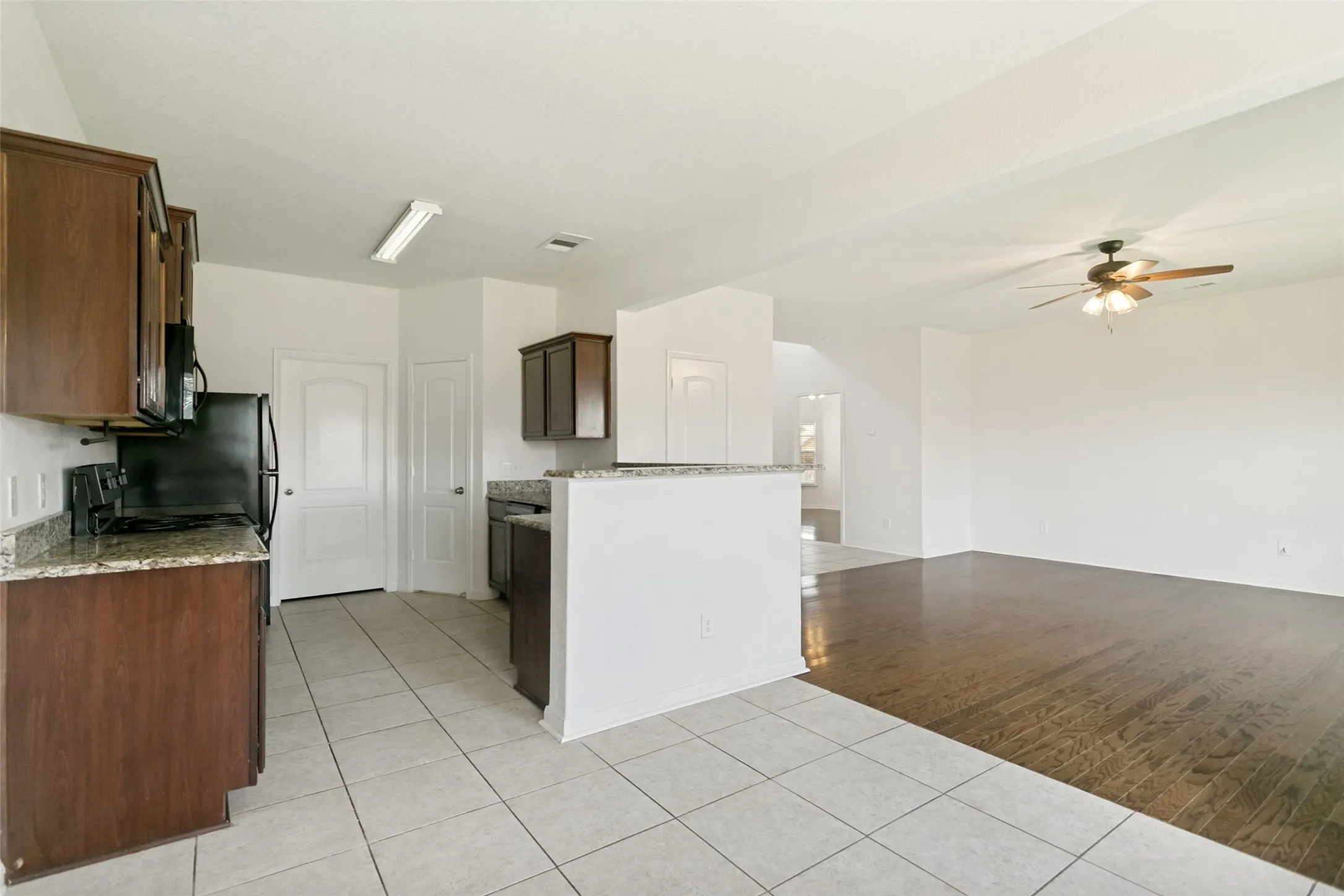 Kitchen featuring tile flooring, granite-style countertops, wood-finish cabinetry, a built-in microwave, and an open concept layout to the adjacent living area with wood-finish flooring and a ceiling fan