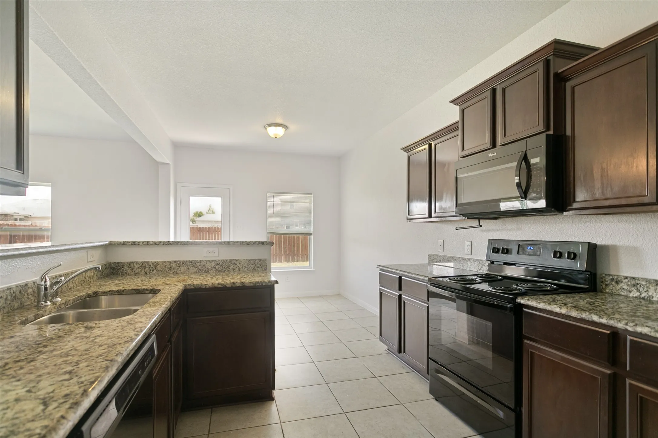 Kitchen with dark wood cabinetry, light speckled countertops, a double basin stainless steel sink, tile flooring, and integrated black appliances