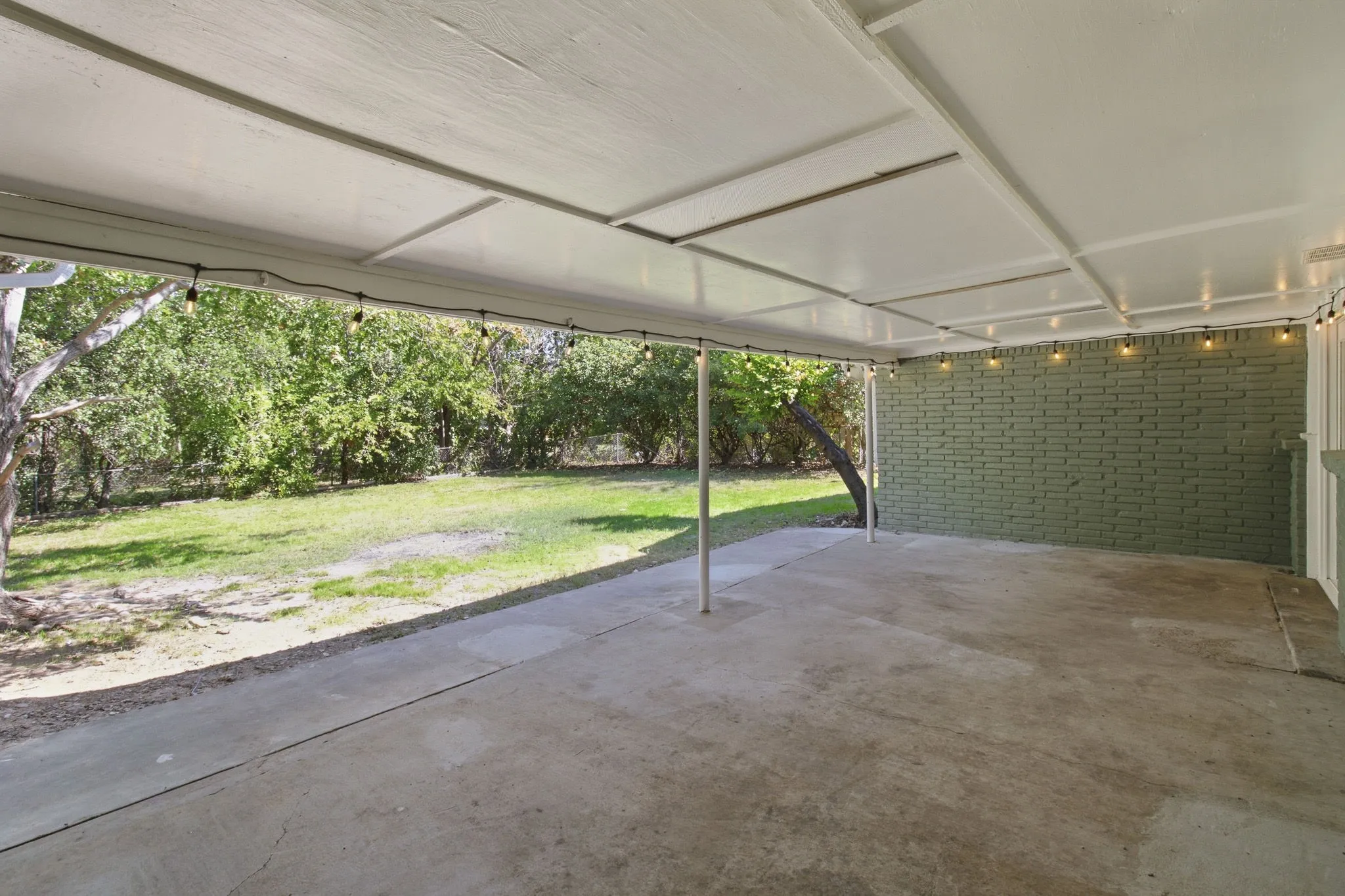 Covered concrete patio featuring string lighting, brick exterior wall, and surrounding lawn area