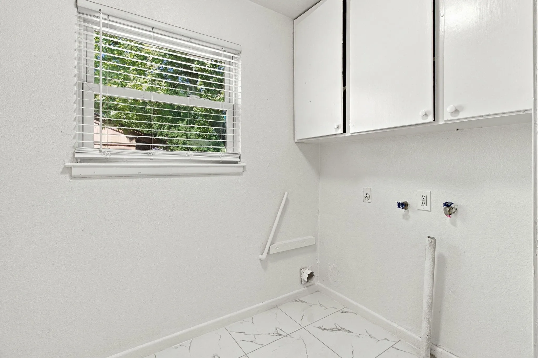 White cabinetry, window with blinds, plumbing hookups, electrical outlets, and tile flooring