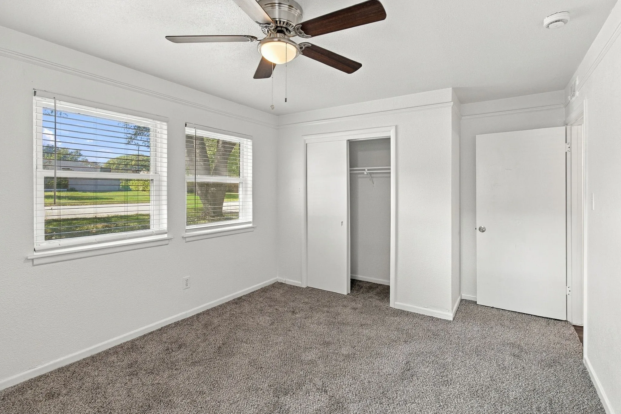 Room featuring white walls, textured carpeting, two windows with blinds, a ceiling fan with light, and a built-in closet with sliding doors