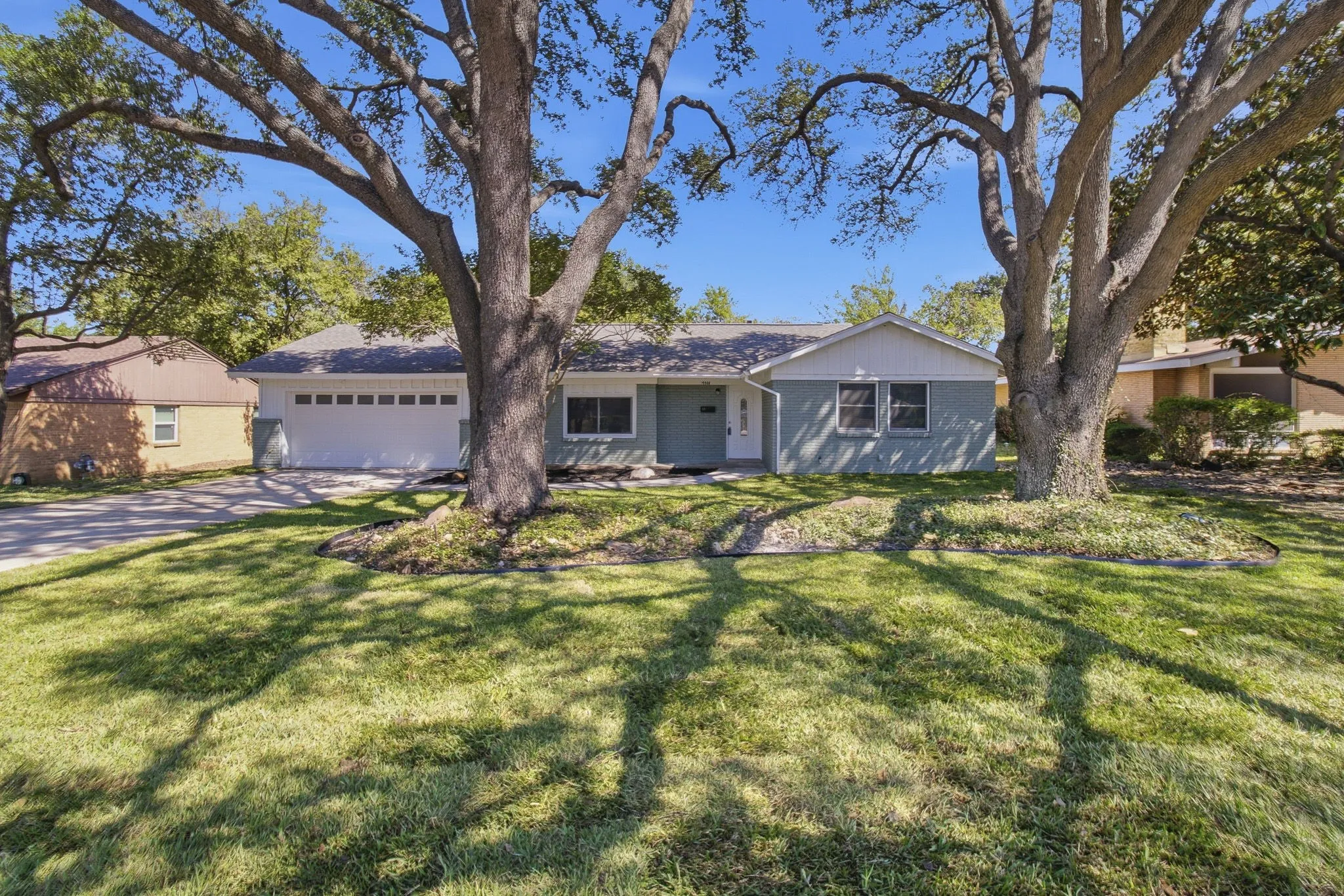 Ranch-style residence featuring a light blue exterior, white trim, attached two-car garage, and established front yard trees