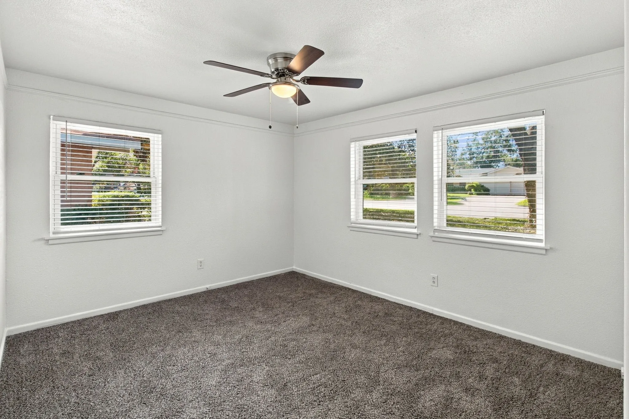 Neutral wall tones, three windows with blinds, gray carpeting, and a ceiling fan with integrated lighting