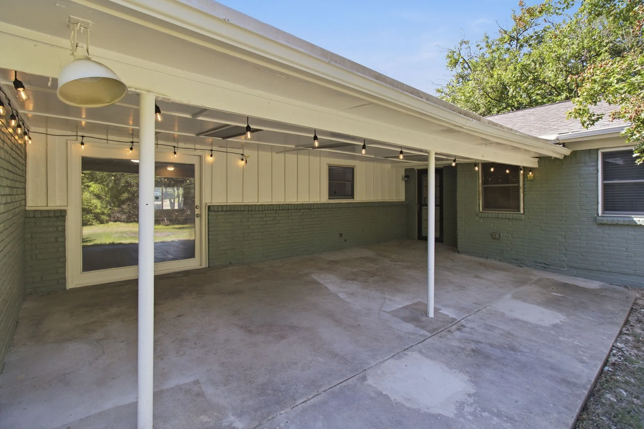 Covered patio with concrete flooring, supported by white posts, featuring string lighting and a central ceiling light fixture